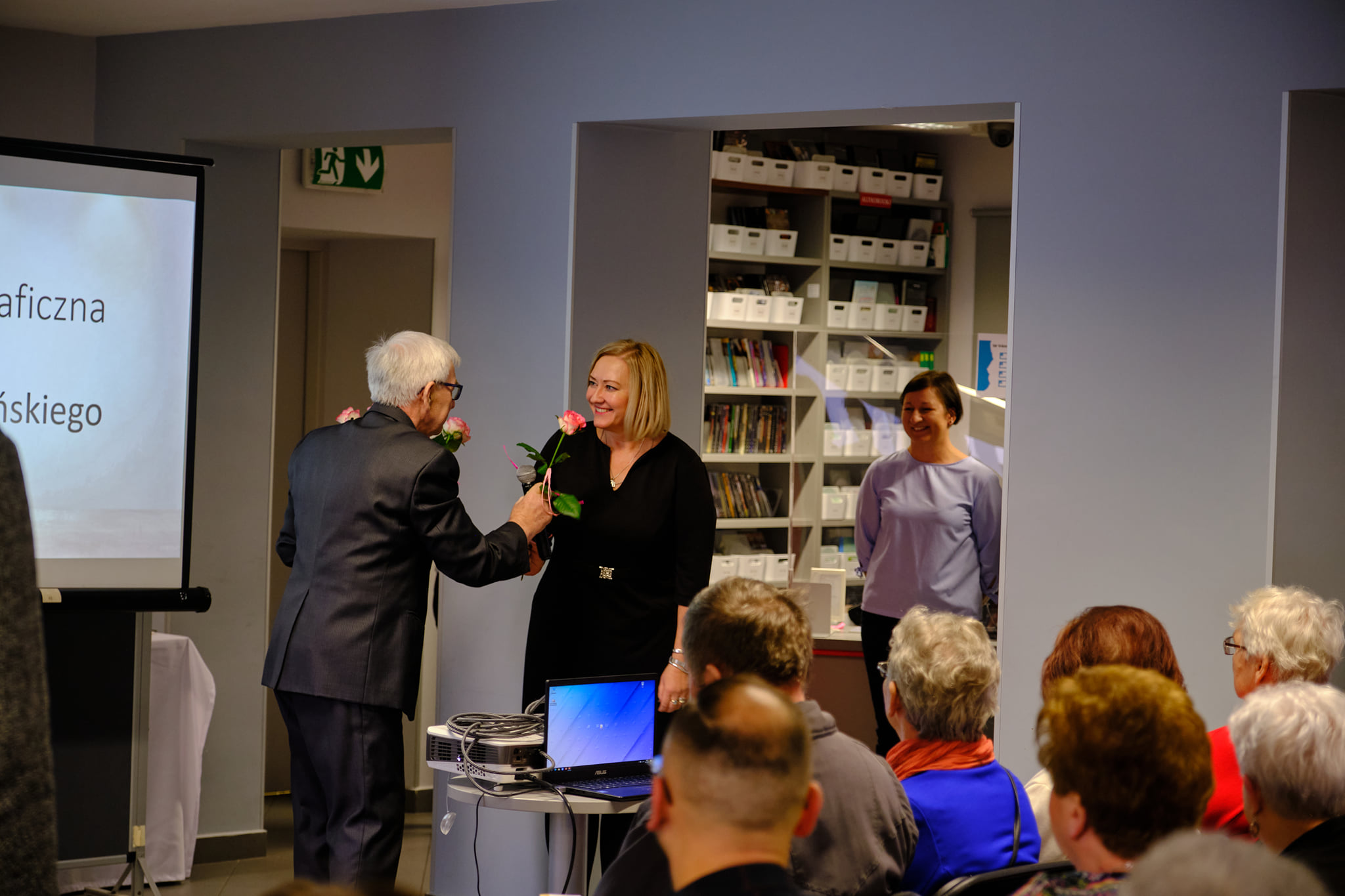 Ryszard Makszyński wręcza kwiaty dyrektorce biblioteki, z przodu zdjęcia widać siedzącą publiczność