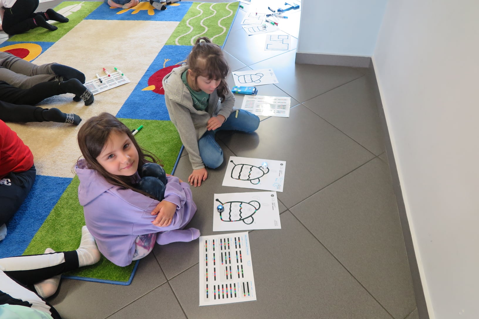 Dwie dziewczynki siedzą na podłodze w tle za nimi widać stopy i nogi innych dzieci leżących na dywanie. Przed dziewczynkami leżą kartki z kolorowymi rysunkami (namalowanymi przez nie trasami dla ozobotów).