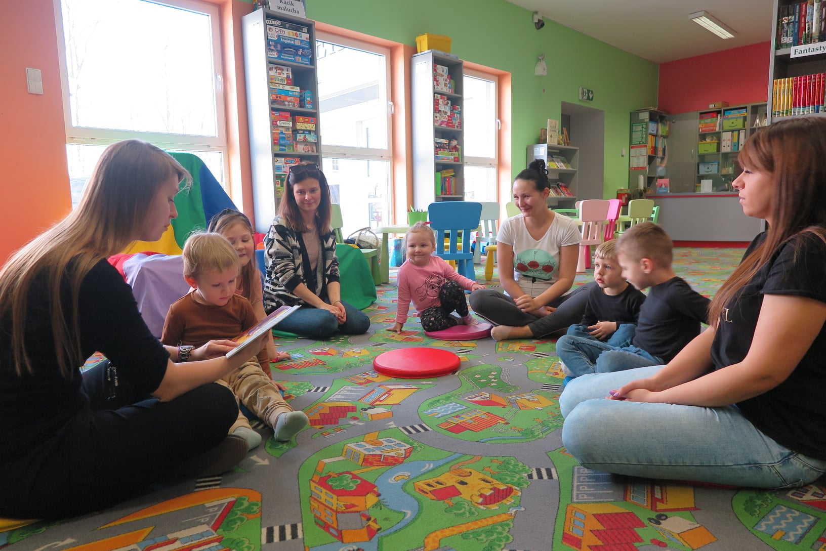 9 osób siedzi na dywanie w oddziale dla dzieci, 5 dzieci, twoje rodziców i bibliotekarka czytająca książkę zgromadzonym
