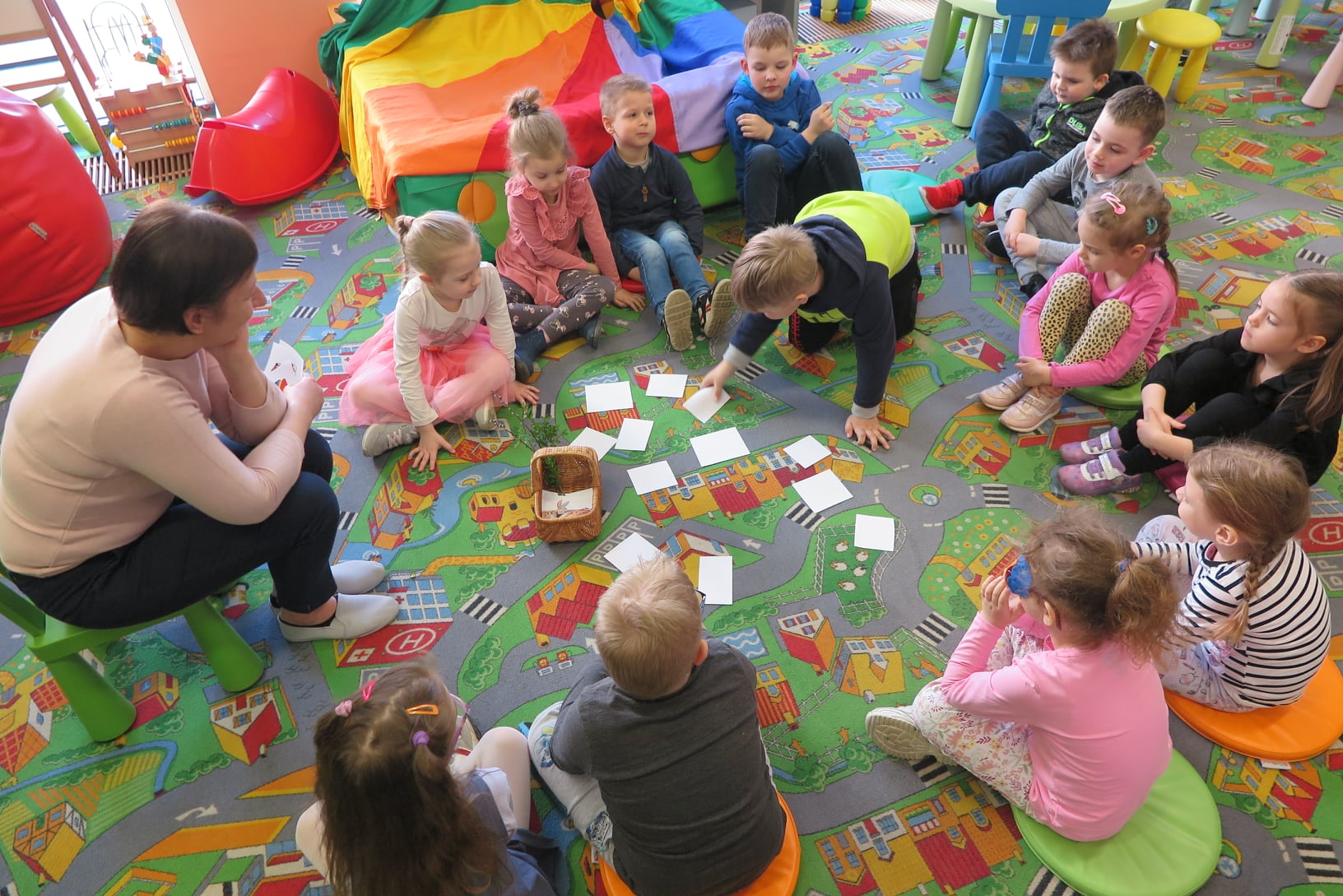 dzieci siedzą na dywanie w kółku w oddziale dla dzieci i wyciągają z wiklinowego koszyka białe karteczki