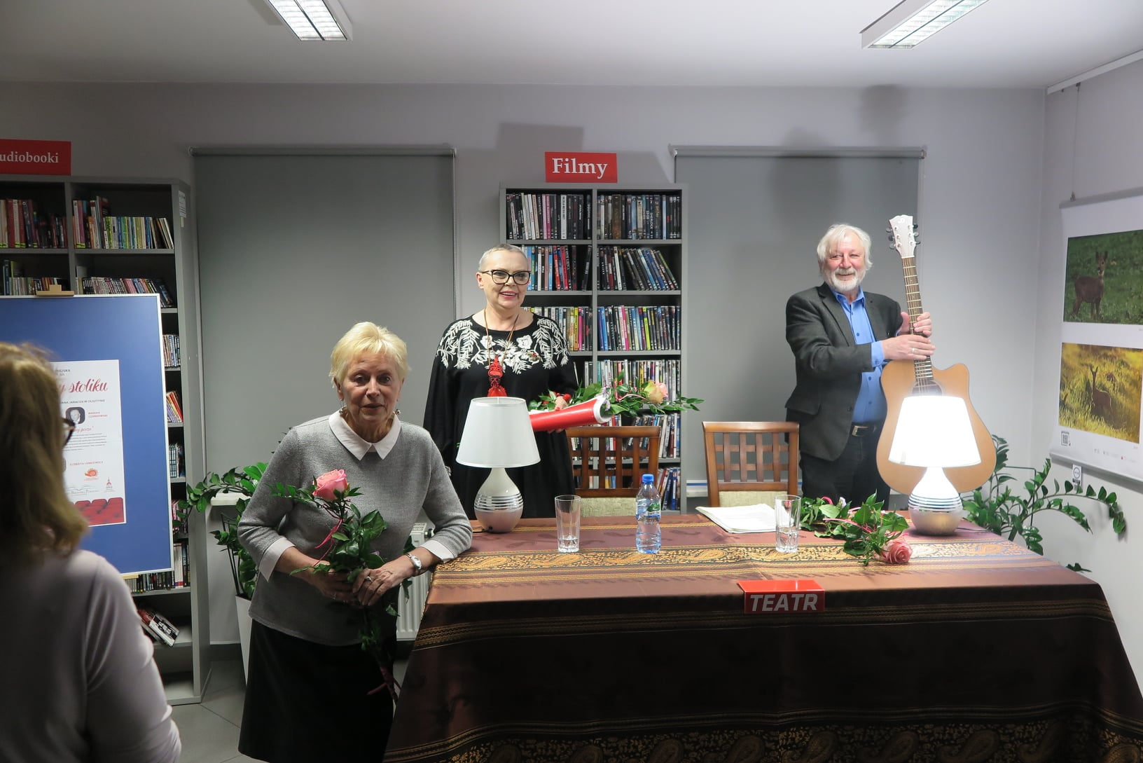 Wiesława Szymańska, Marian Czarkowski i Elżbieta Lenkiewicz stoją przy stoliku z kwiatami, Marian trzyma w ręku gitarę