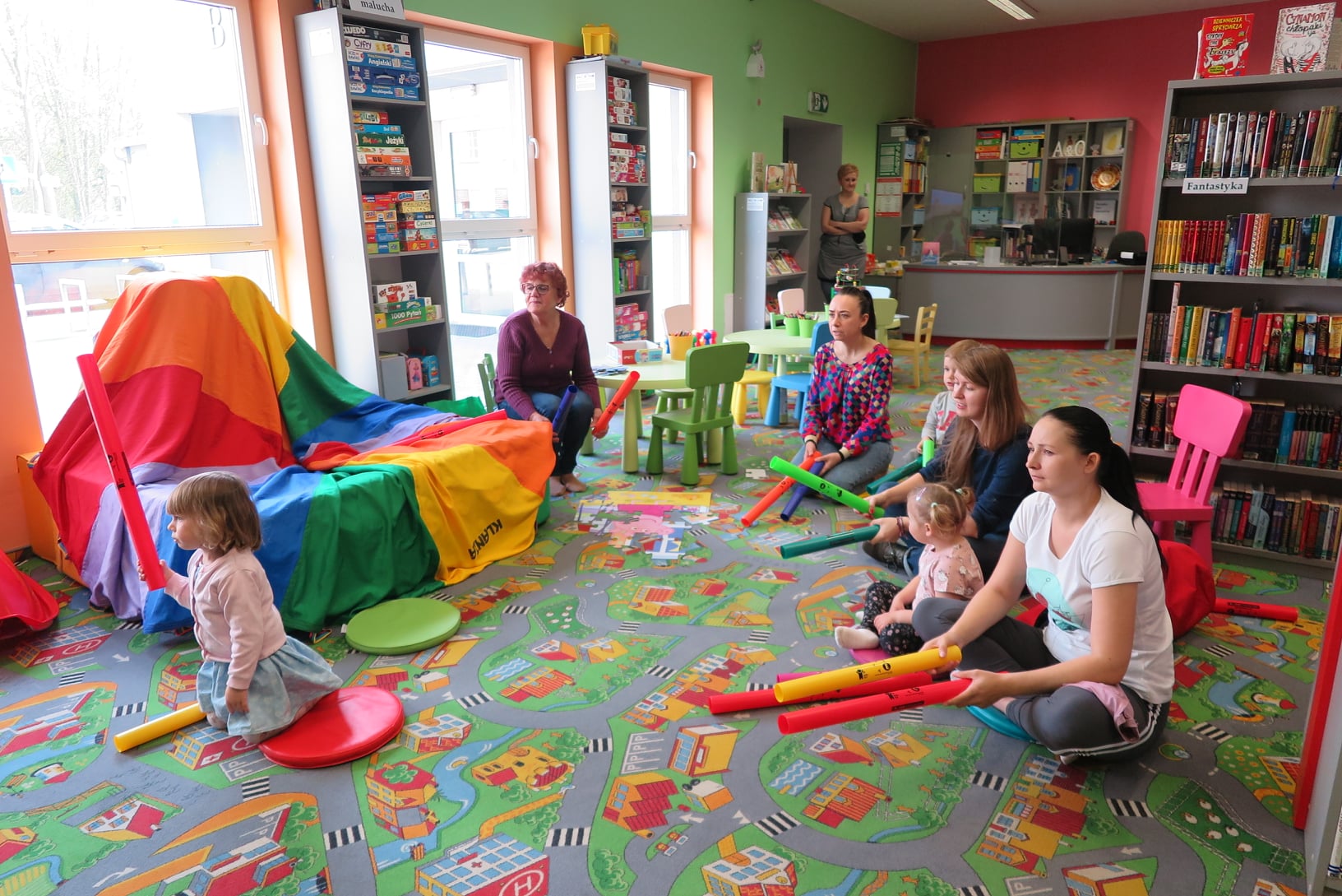 dzieci z rodzicami siedzą na dywanie w oddziale dla dzieci i grają na bum bum rurkach