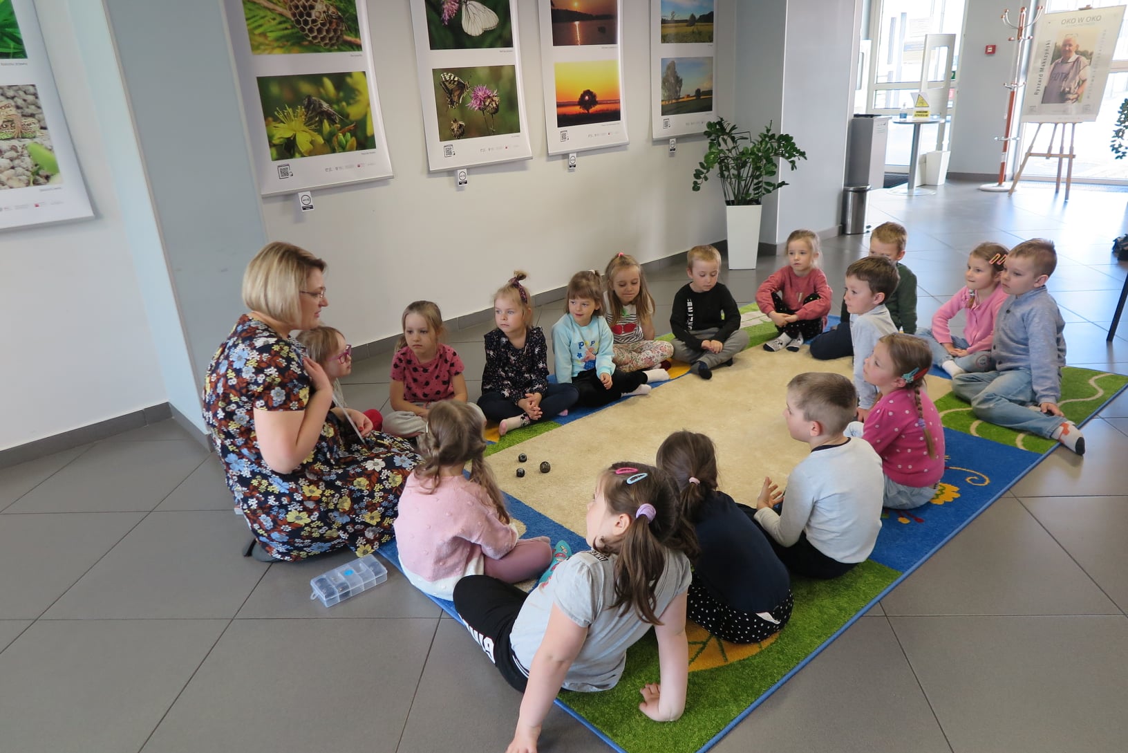 grupa dzieci siedzi na dywanie w mediatece, bibliotekarka pokazuje im kolorowe kody do sterowanie ozobota