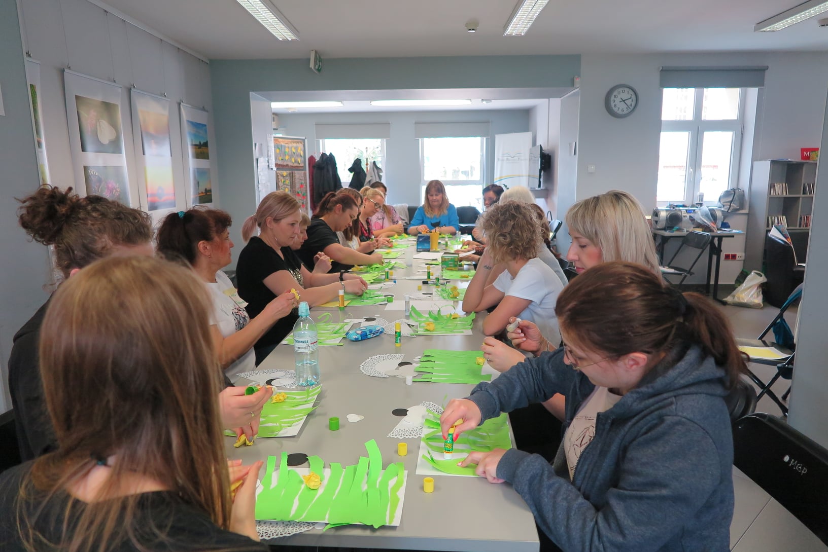 grupa kobiet siedzi przy długim stoliku i wyklejają z zielonego papieru pracę plastyczną