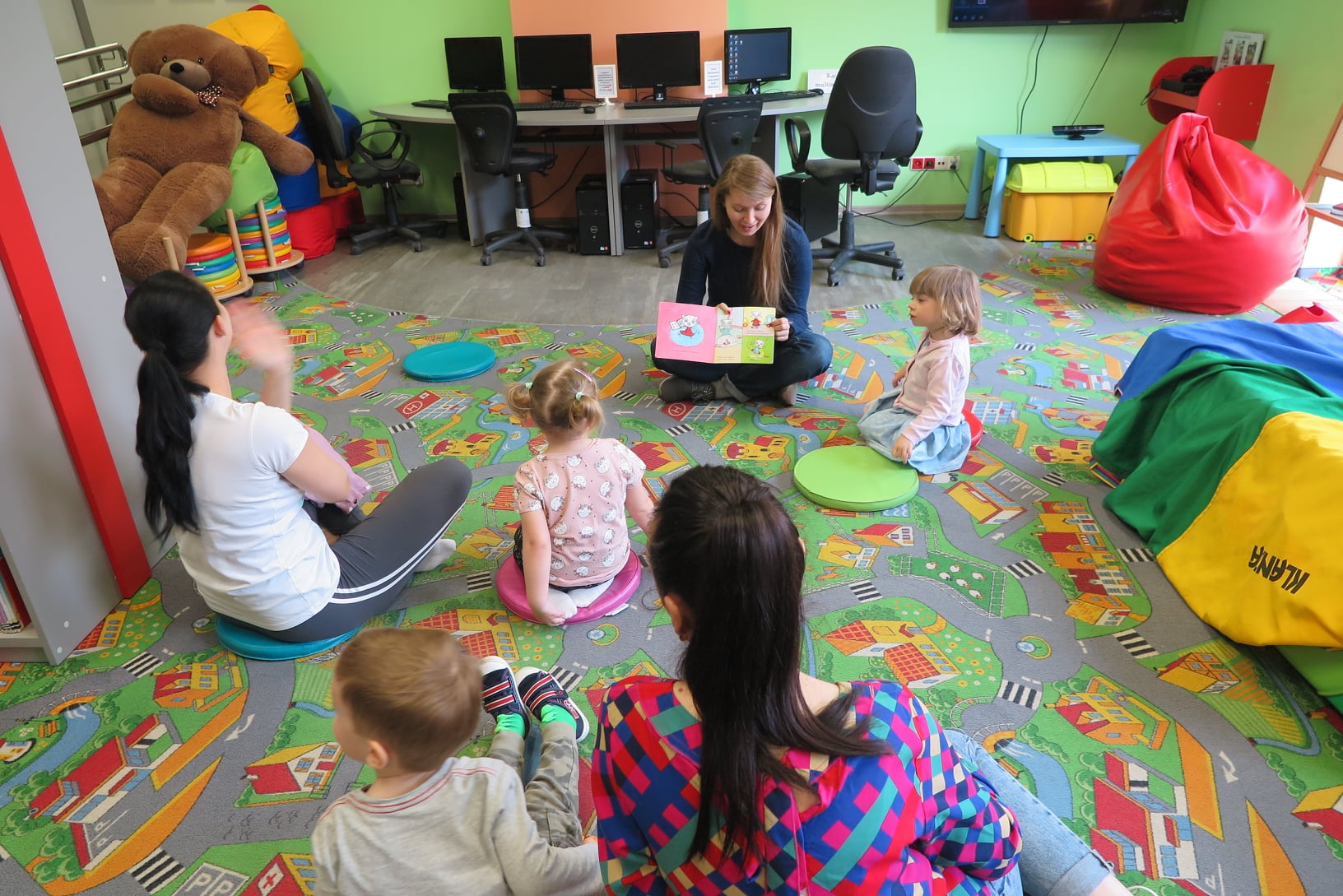 dzieci z rodzicami siedzą na dywanie w oddziale dla dzieci i słuchają opowieści czytanej przez bibliotekarką