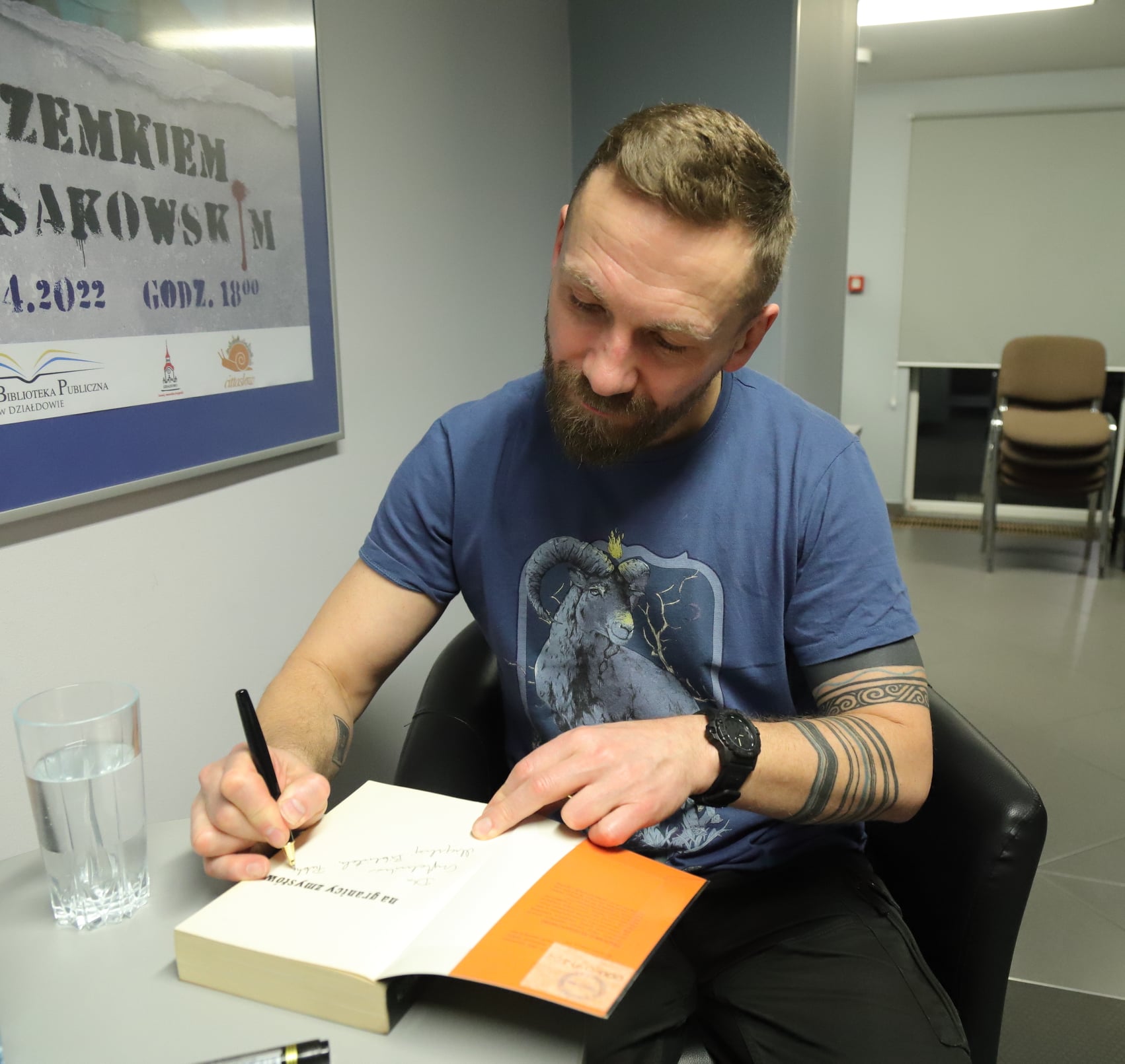 Przemek Kossakowski  siedzi w fotelu i składa autograf na okładce książki