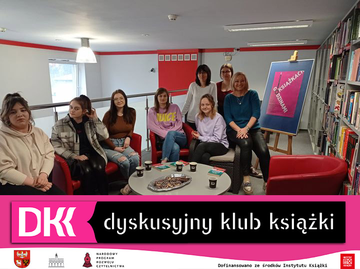8 kobiet siedzi przy okrągłym stole na antresoli biblioteki, obok stoi sztaluga z plakatem DKK