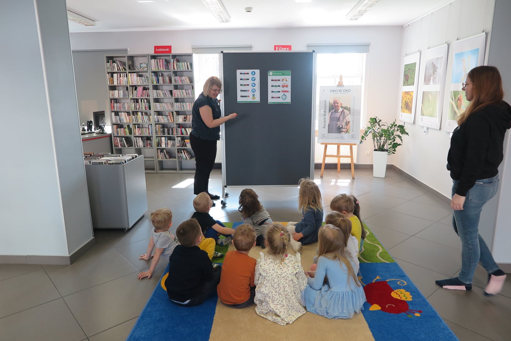 wnętrze mediateki, na dywanie siedzą dzieci, przyglądają się tablicy na której wiszą kody programowania ozobota, obok tablicy stoi bibliotekarka, po prawej stroni zdjęcia stoi opiekunka dzieci