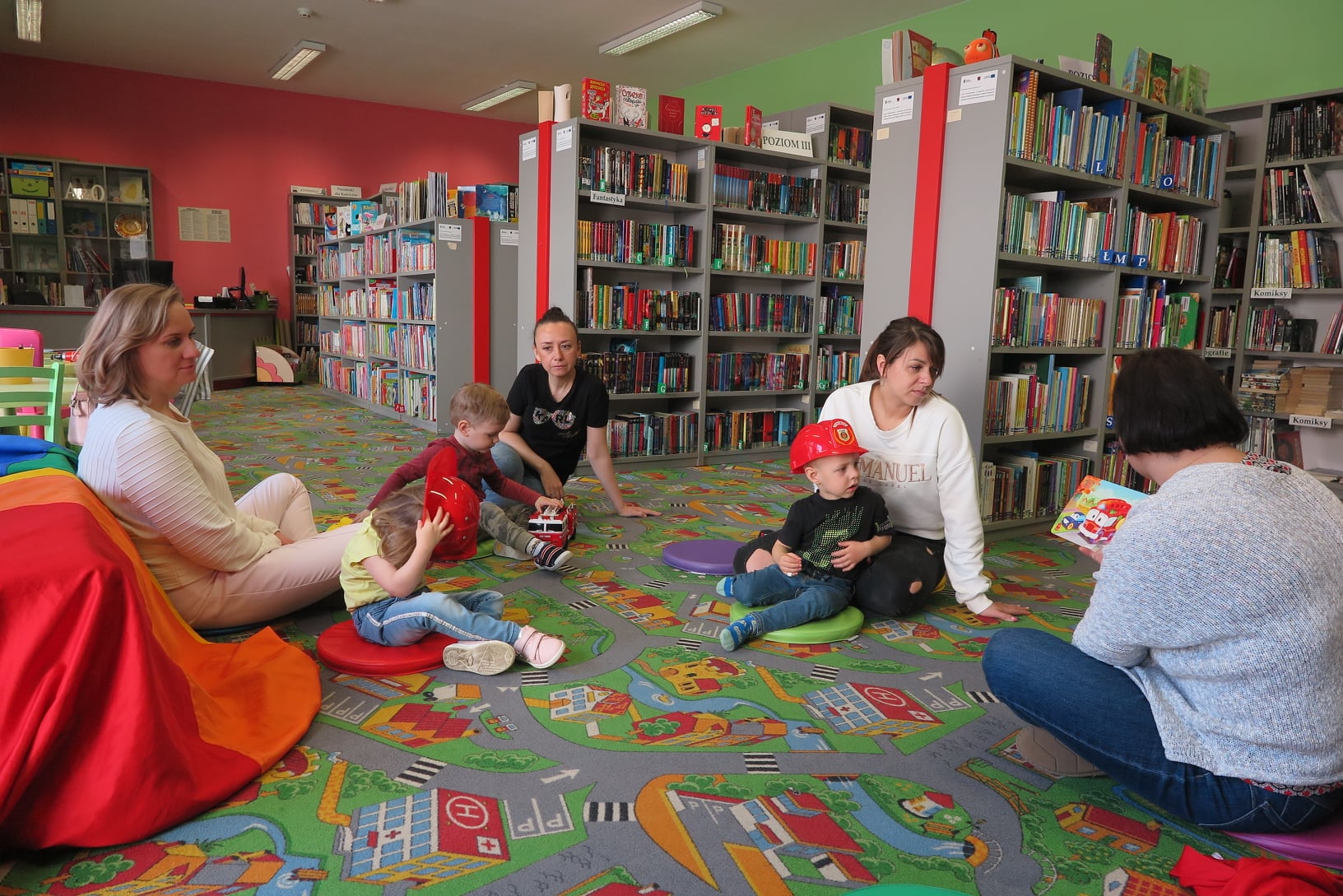 dzieci z opiekunami  siedzą na podłodze w oddziale dla dzieci, bibliotekarka czyta książkę, dzieci siedzą na kolorowych krążkach