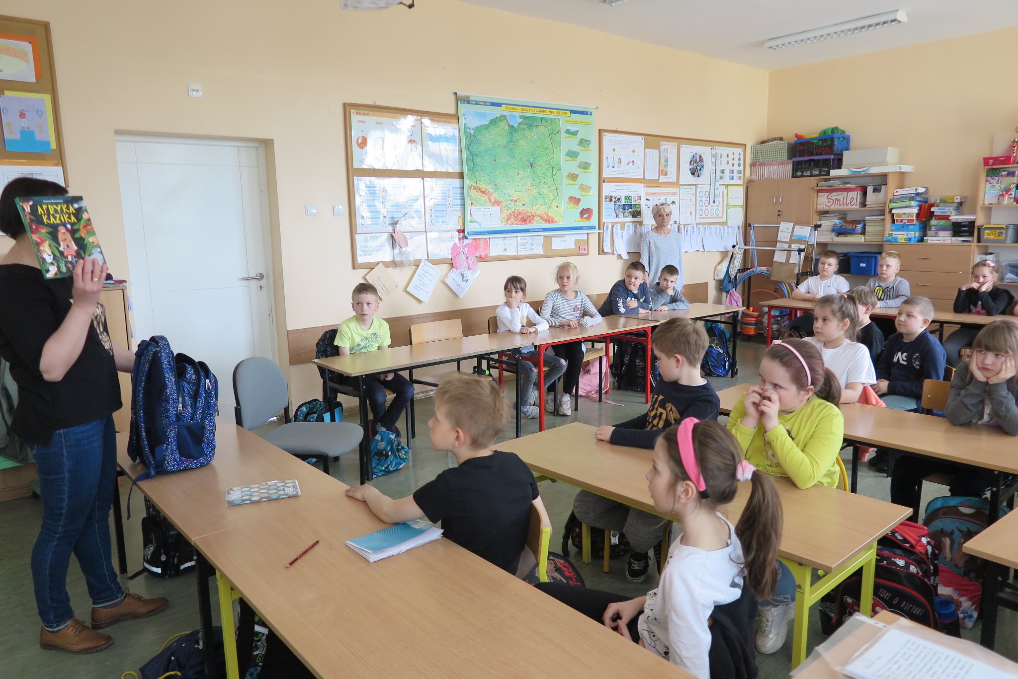 grupowe zdjęcie dzieci, nauczycielki i ze szkoły podstawowej w sali lekcyjnej, dzieci siedzą w swoich ławkach szkolnych, bibliotekarka trzyma w ręku książkę wyjętą z biblio-plecaka