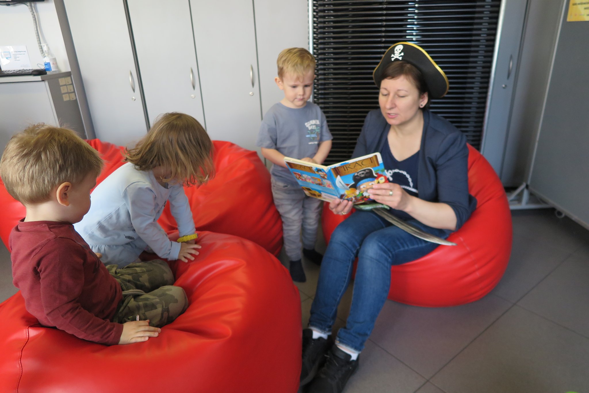 bibliotekarka  z dziećmi siedzą na pufach i czyta im książkę