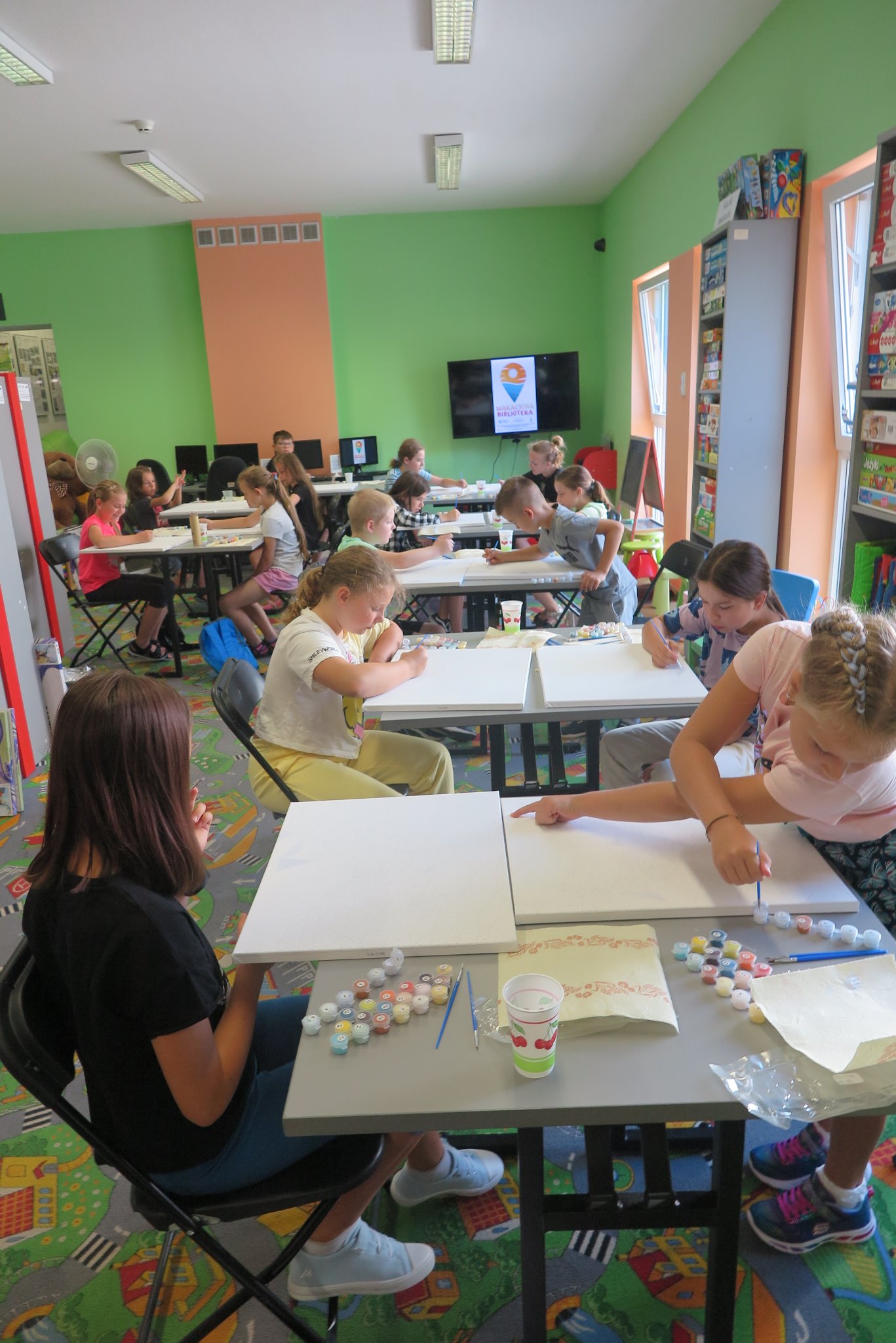 dzieci siedzą przy stolikach w oddziale dla dzieci, na stolikach leżą zestawy do malowania obrazów