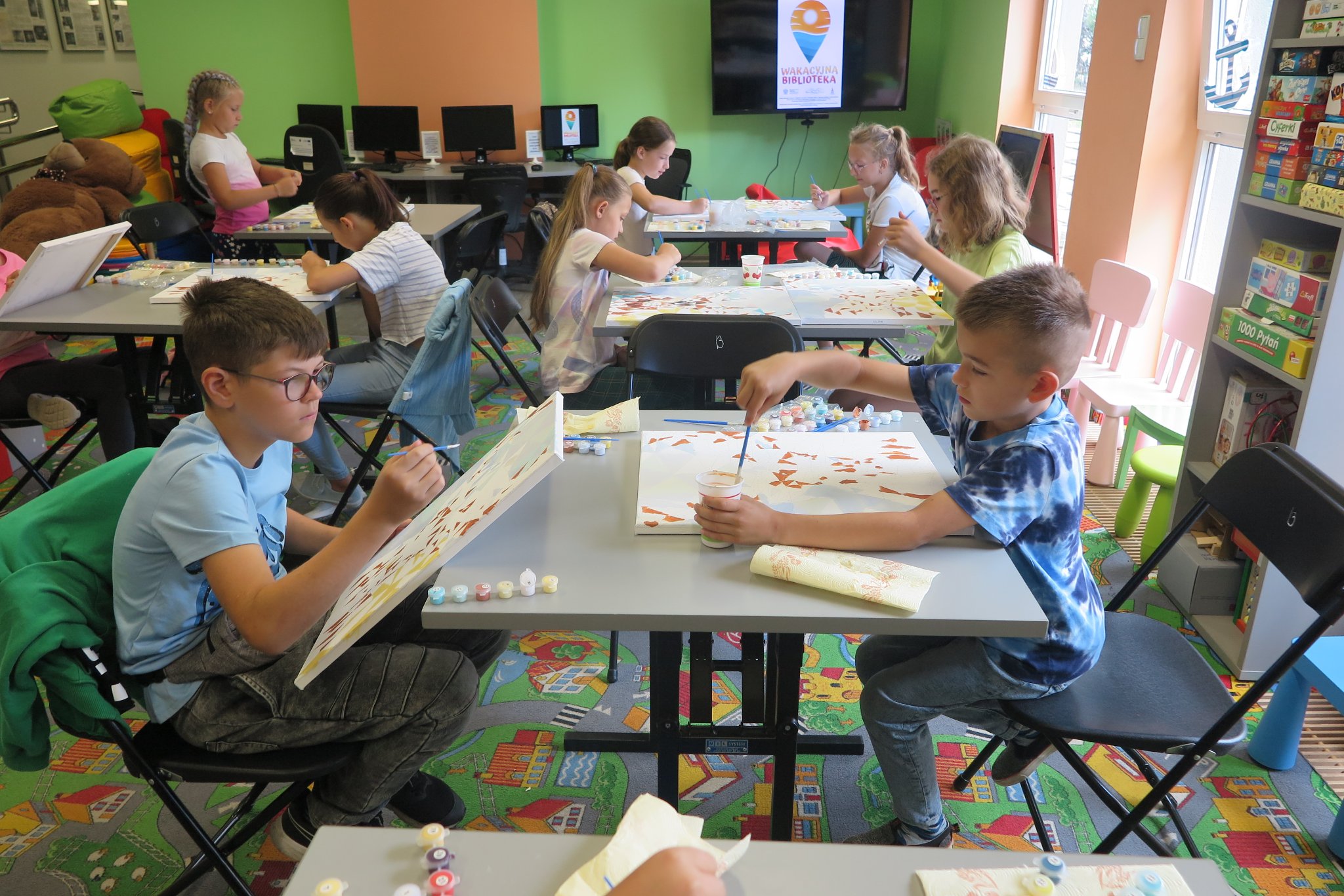dzieci siedzą przy stolikach w oddziale dla dzieci, na stolikach leżą zestawy do malowania obrazów, dzieci malują obrazy