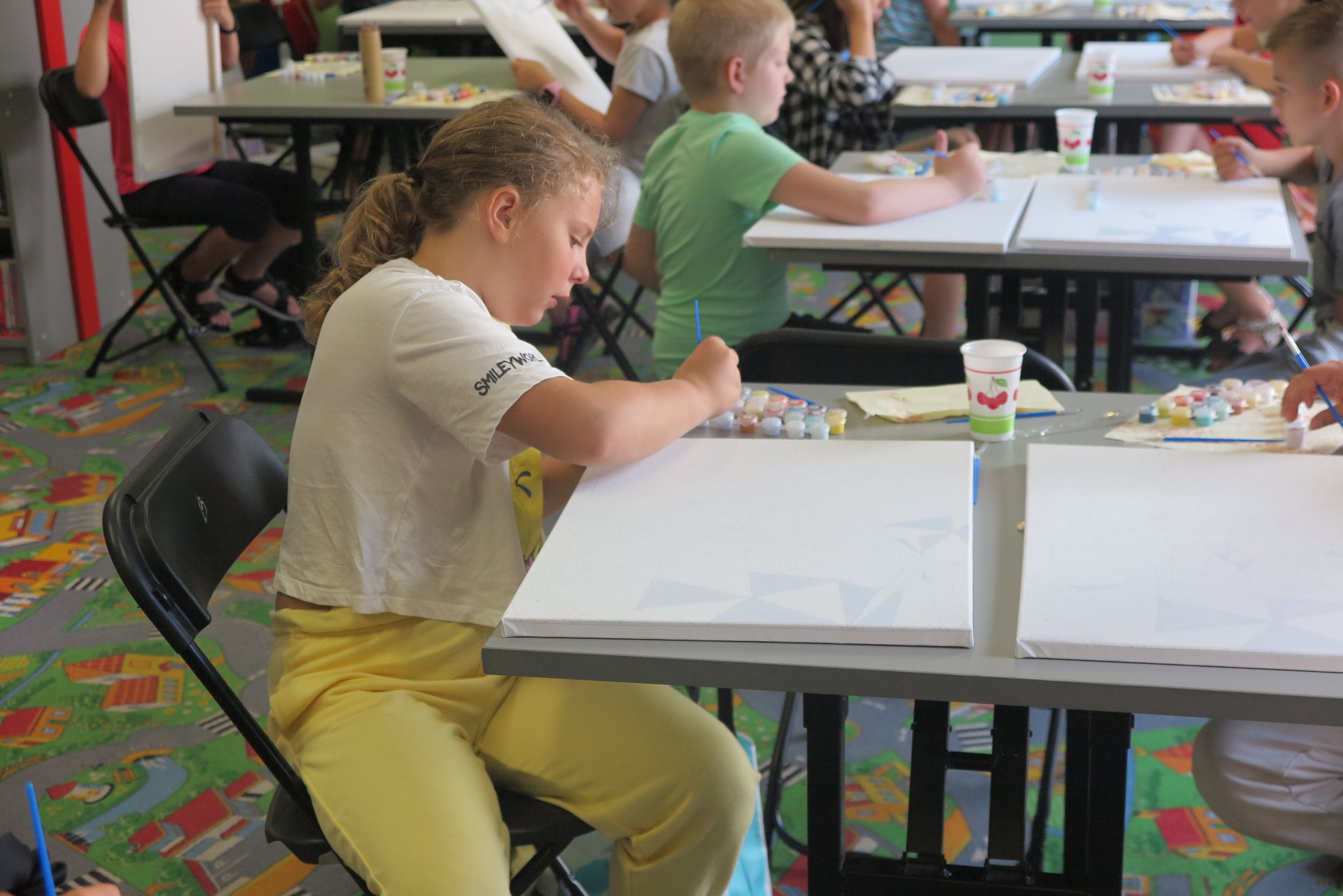 dzieci siedzą przy stolikach w oddziale dla dzieci, na stolikach leżą zestawy do malowania obrazów, dzieci malują obrazy