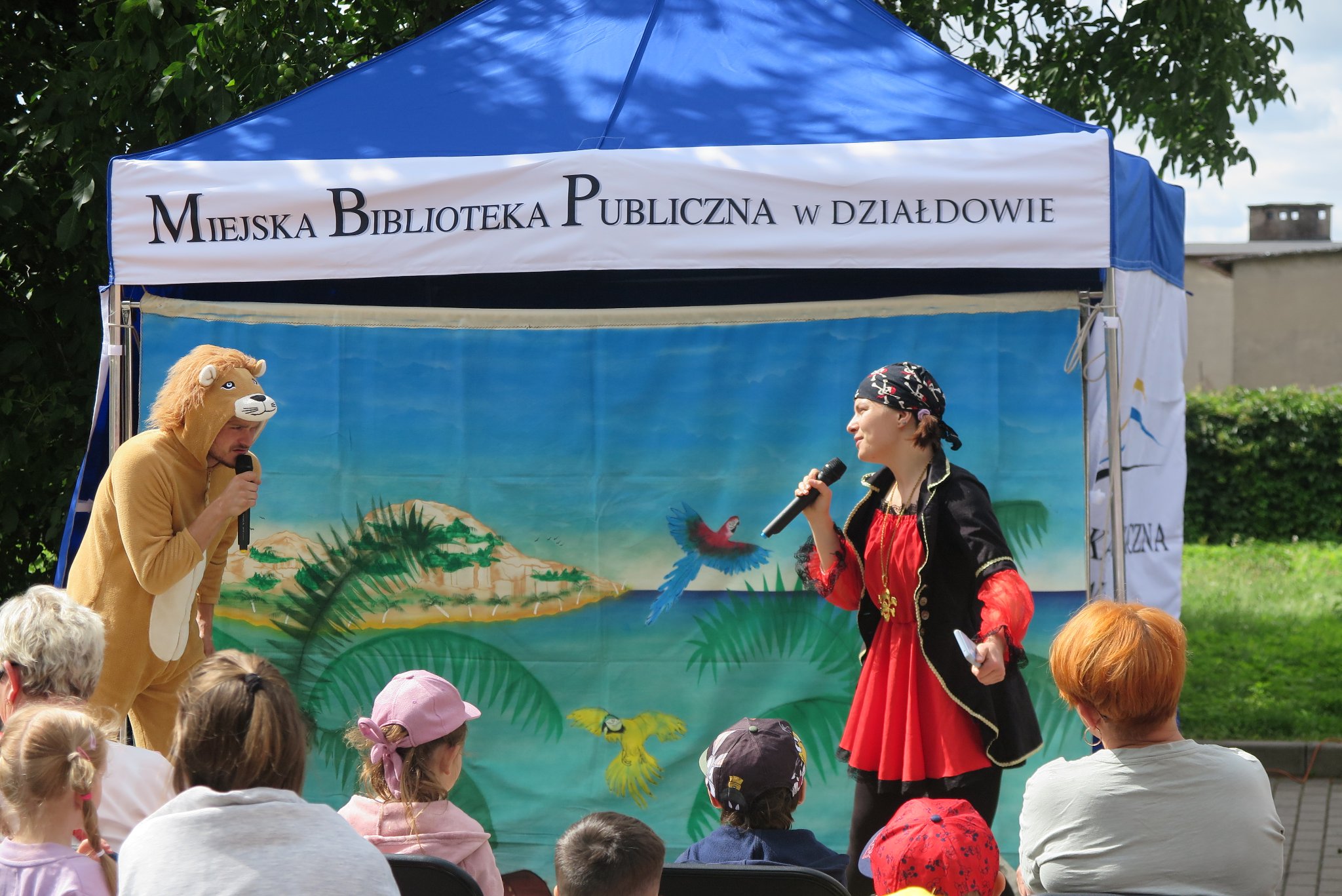 dwoje aktorów (lew i pirat) teatru Maska  na tle kurtyny dekoracji  z wyspą i palmami, przed nimi siedzi publiczność
