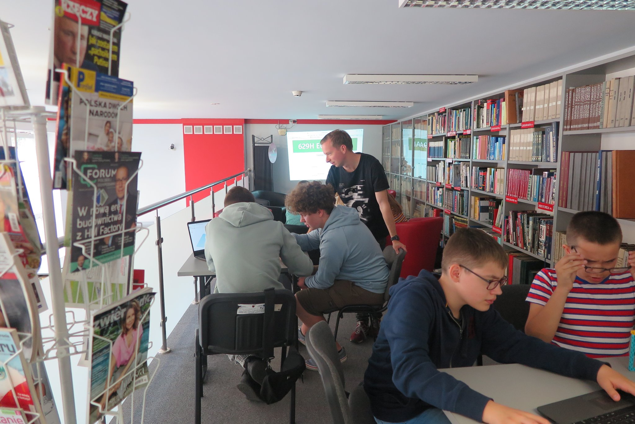 antresola biblioteki, trener i uczestnicy warsztatów siedzą przy stolikach  z na których leżą laptopy, w tle baner Klub Młodego Programisty, na ścianie wyświetlany jest obraz z rzutnika