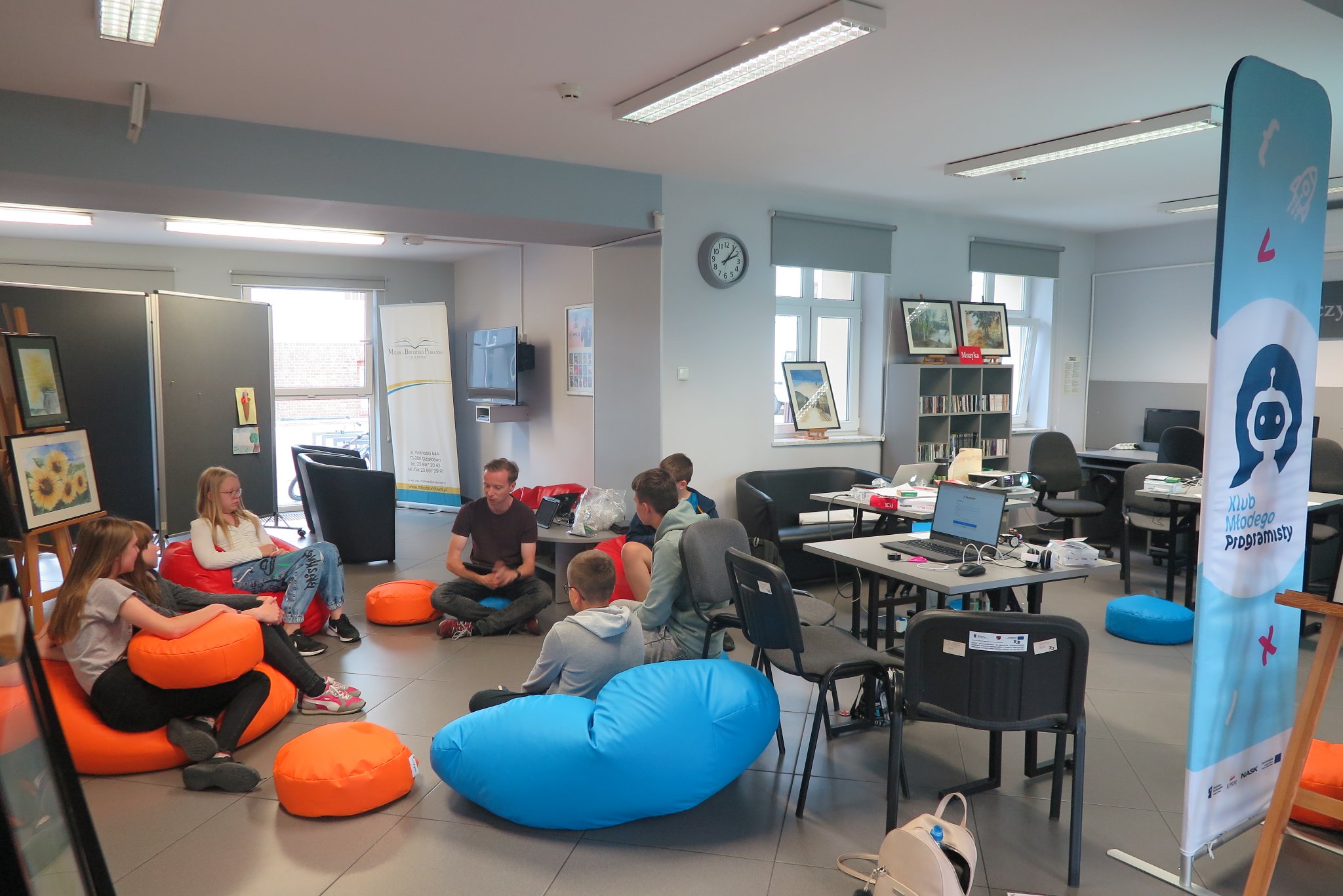 wnętrze mediateki, trener i uczestnicy warsztatów siedzą na kolorowych pufach, obok na stolikach  leżą laptopy, z prawej strony stoi baner Klub Młodego Programisty