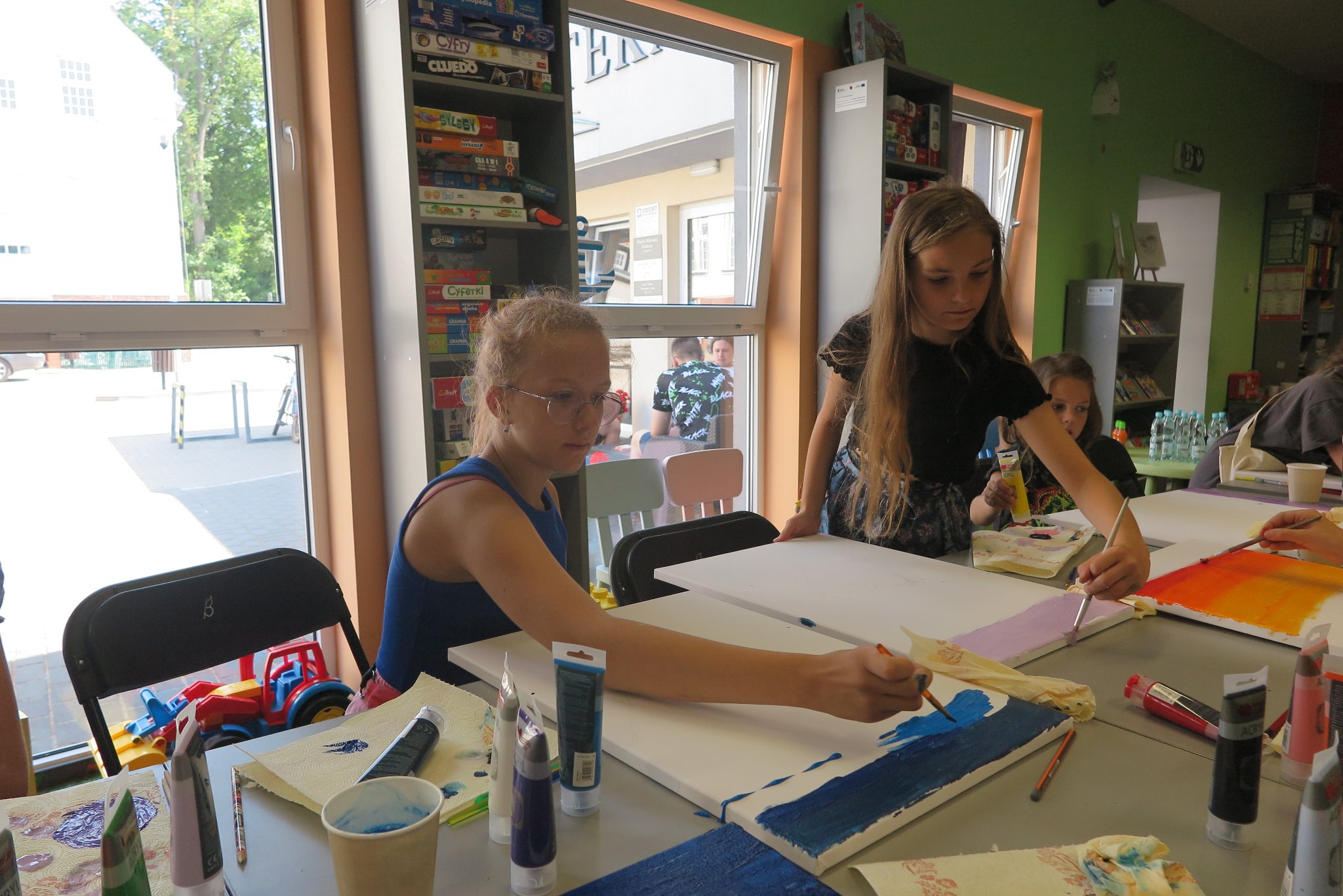 uczestnicy warsztatów malowania na płótnie siedzą na krzesłach przy stoliku i malują obrazy leżące na blacie stołu