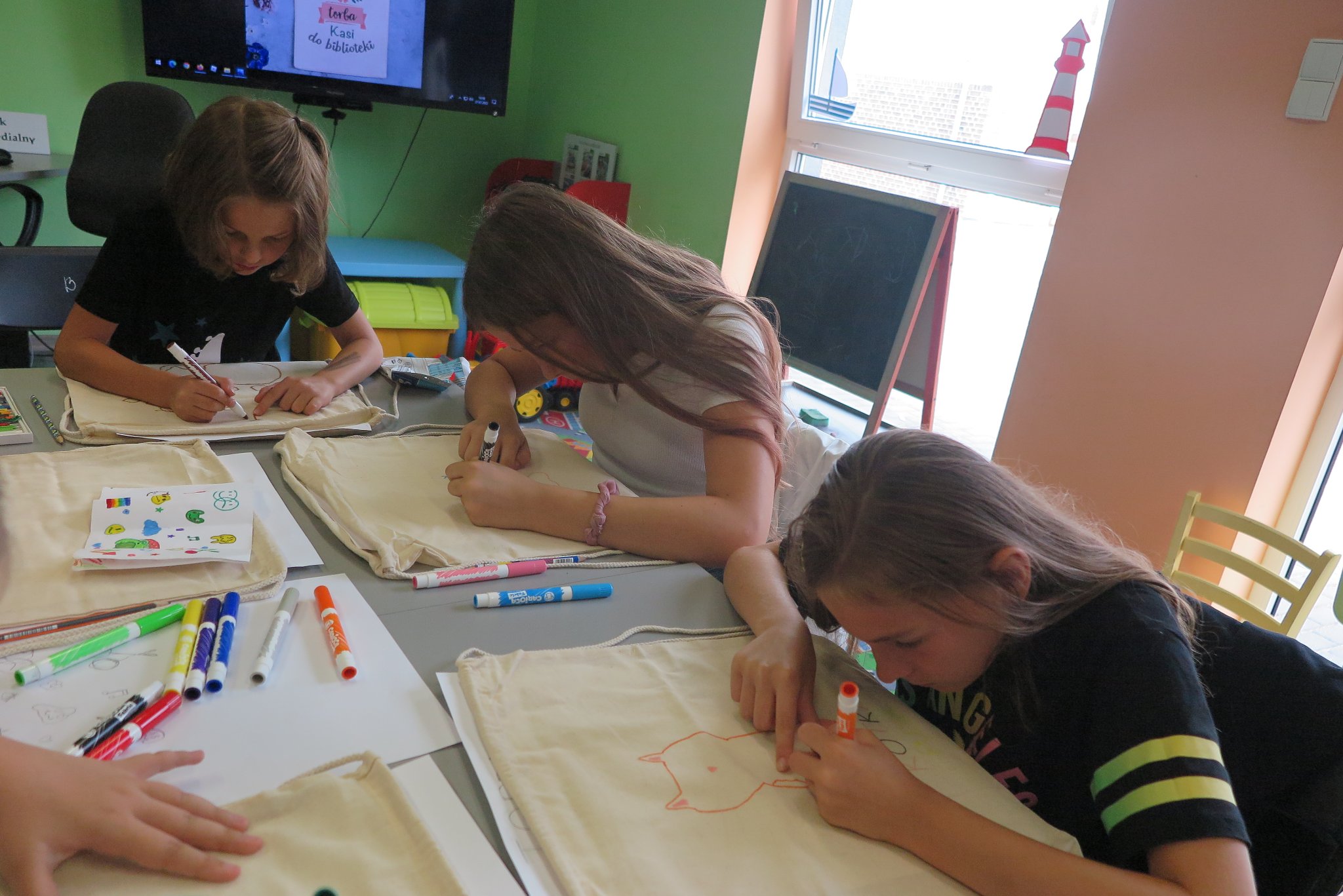 uczestnicy zajęci plastycznych z malowania toreb siedzie przy długim stole na którym leżą bawełniane białe torby i mazaki do pisania po tkaninie, dzieci malują torby