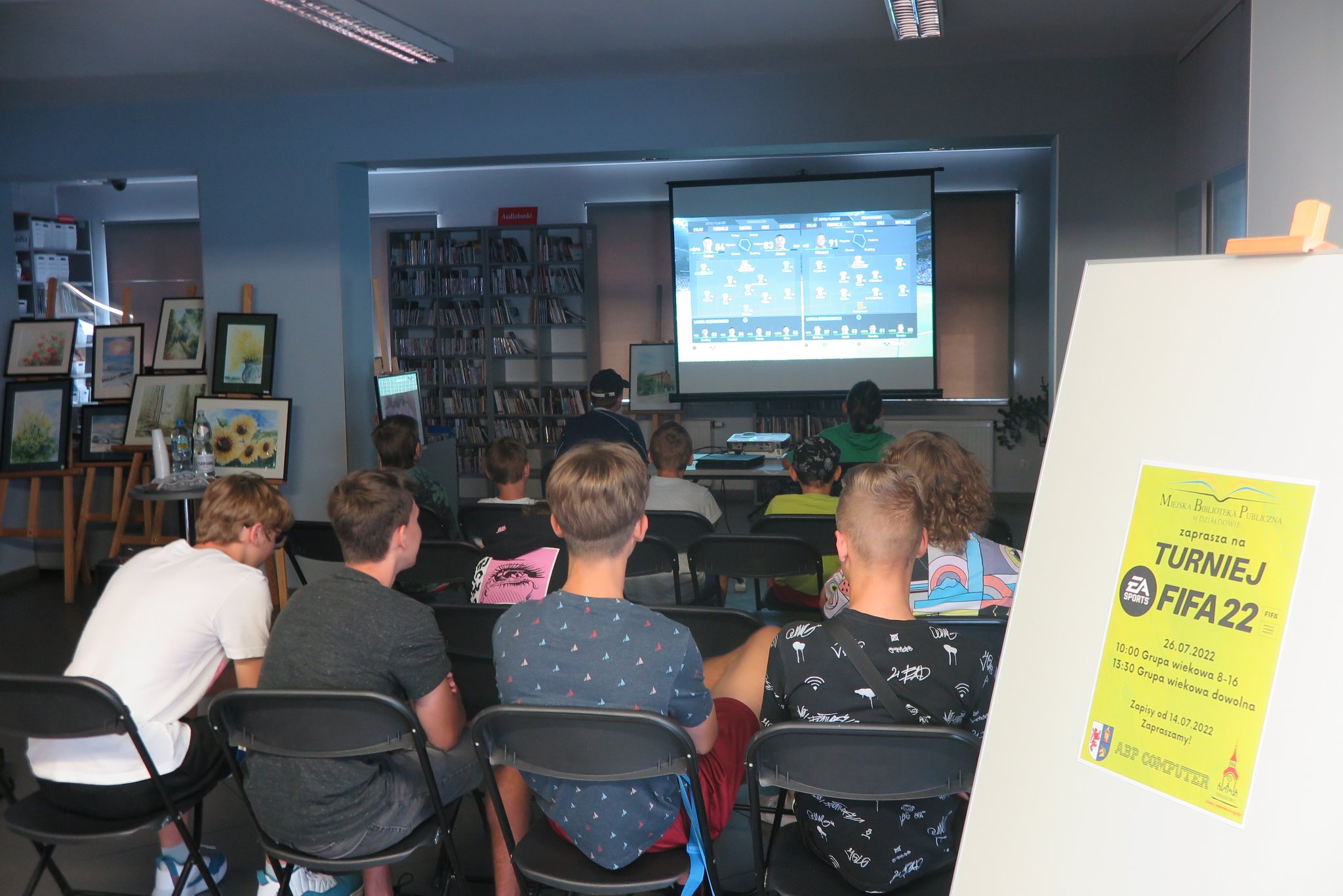 zdjęcie wykonane z tyłu na którym widać publiczność oraz ekran projektora, z prawej na sztaludze stoi plakat turnieju Fifa 22