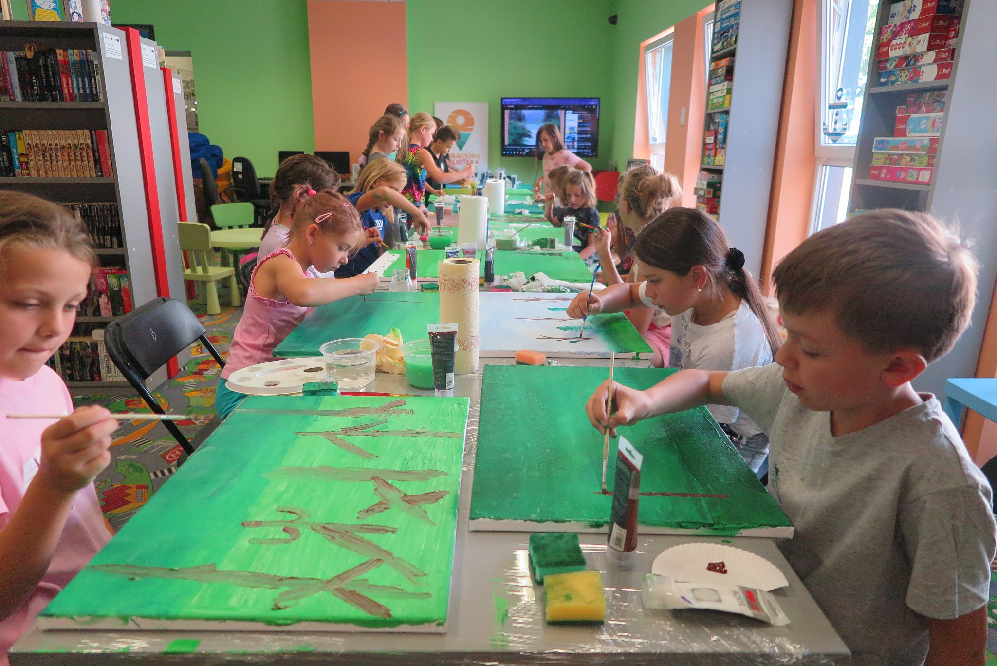 uczestnicy warsztatów malowania na płótnie siedzą na krzesłach przy stoliku i malują obrazy leżące na blacie stołu