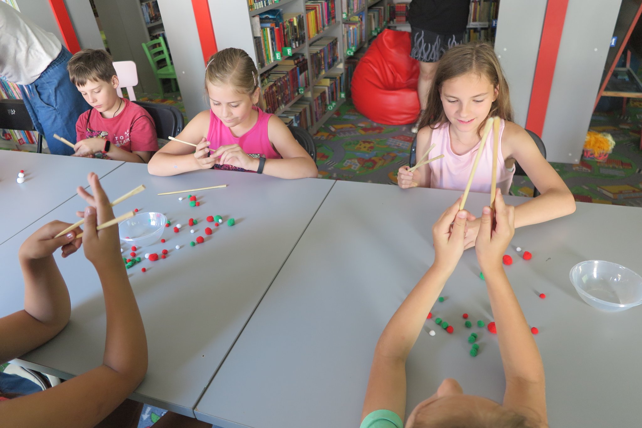 uczestnicy warsztatów podróżniczych z Anną Jaklewicz uczą się posługiwać pałeczkami do jedzenia, ćwiczą na kolorowych kulkach, siedzą przy stole w oddziale dla dzieci