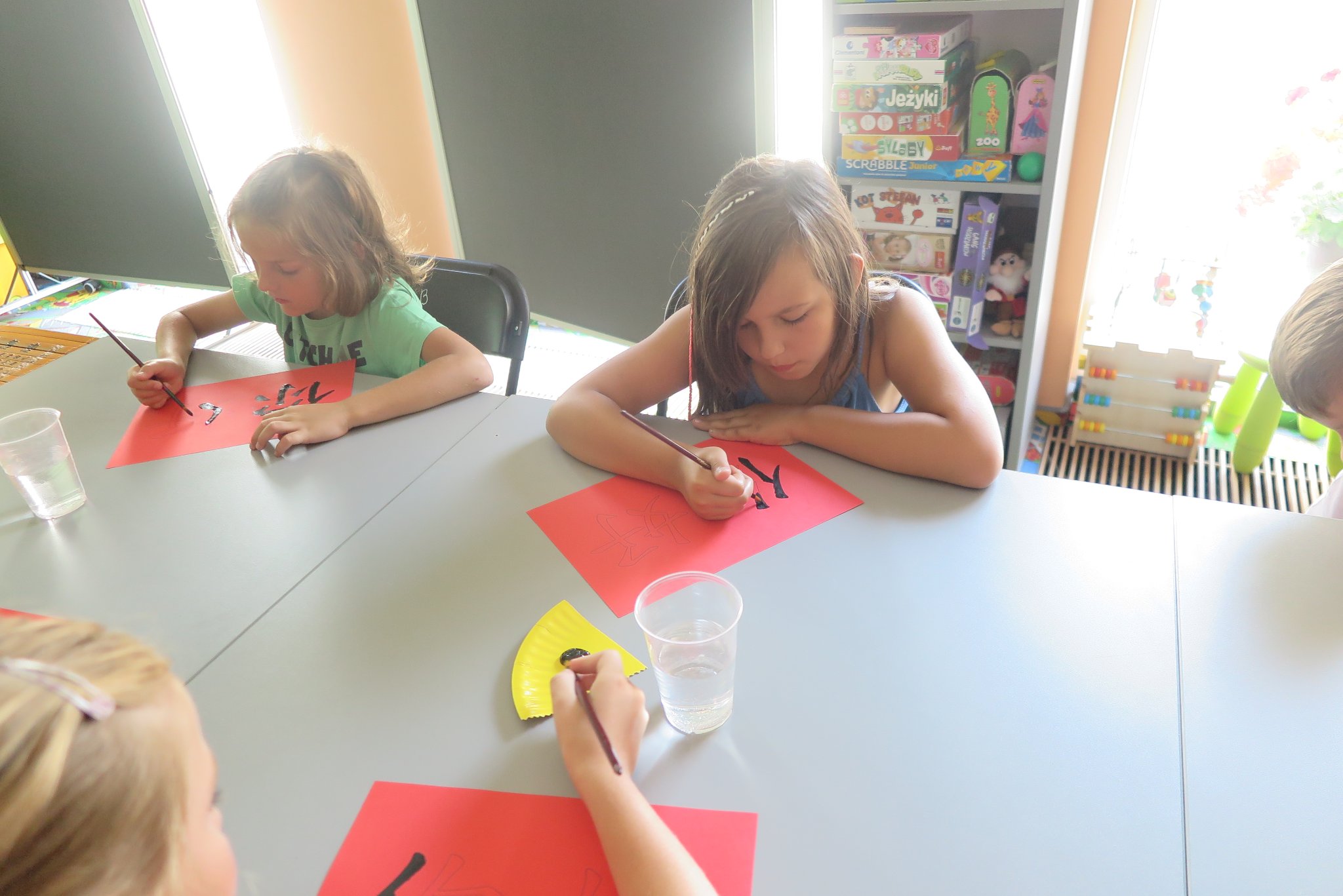uczestnicy warsztatów podróżniczych z Anną Jaklewicz uczą się pisać chińskie symbole na czerwonych kartkach, siedzą przy stole w oddziale dla dzieci