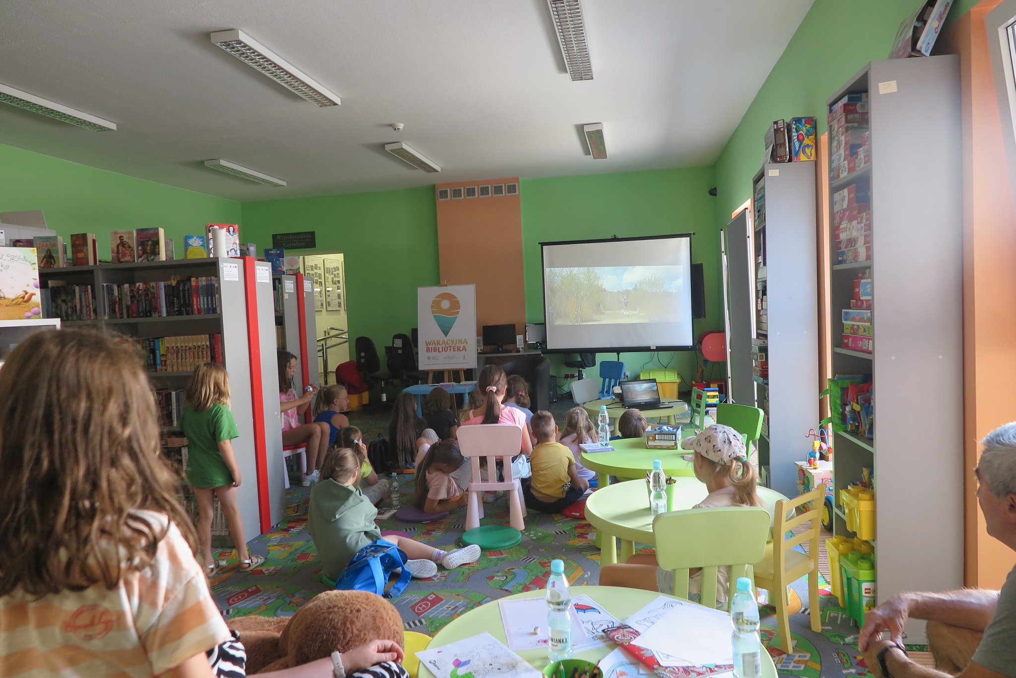 uczestnicy Wakacyjnego Klubu Książki oglądają na ekranie projektora ekranizację przeczytanej przez bibliotekarkę książki, siedzą na podłodze na kolorowych okrągłych poduszkach