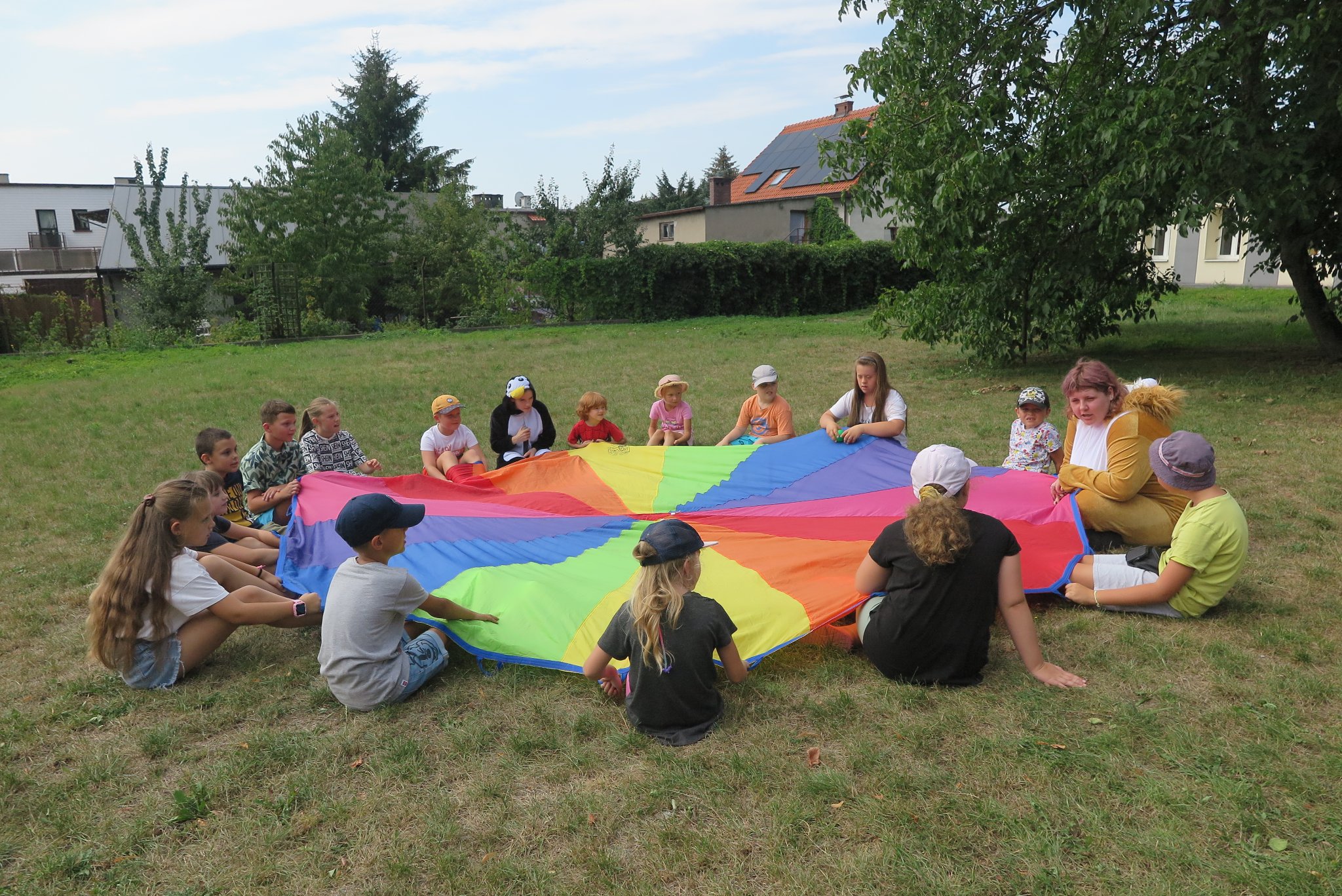 dzieci siedzą na trawie po okręgu, trzymają w rękach rozciągnięty "spadochron"