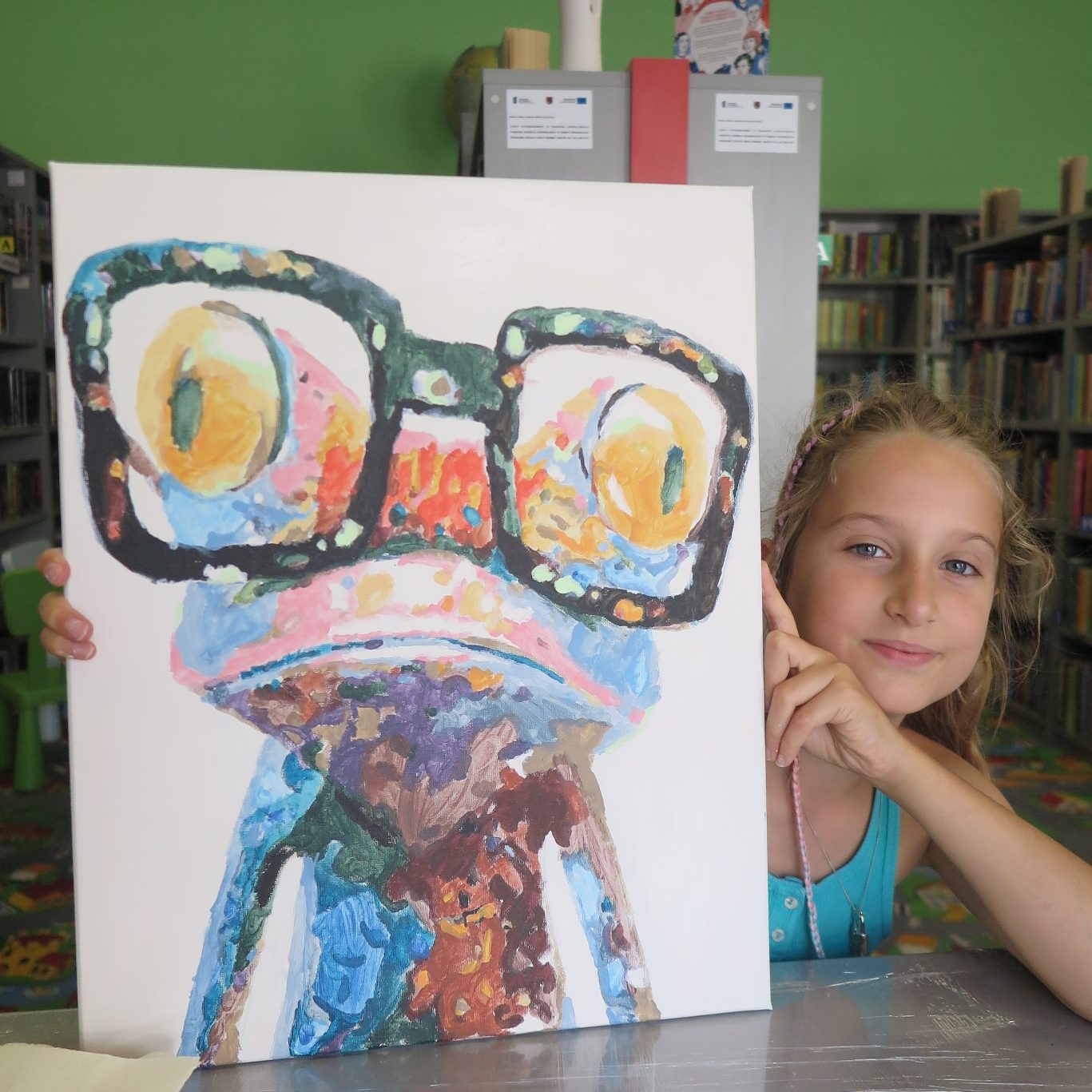 dziewczynka siedzi przy stole na którym trzyma namalowany na zajęciach obraz żaby w okularach