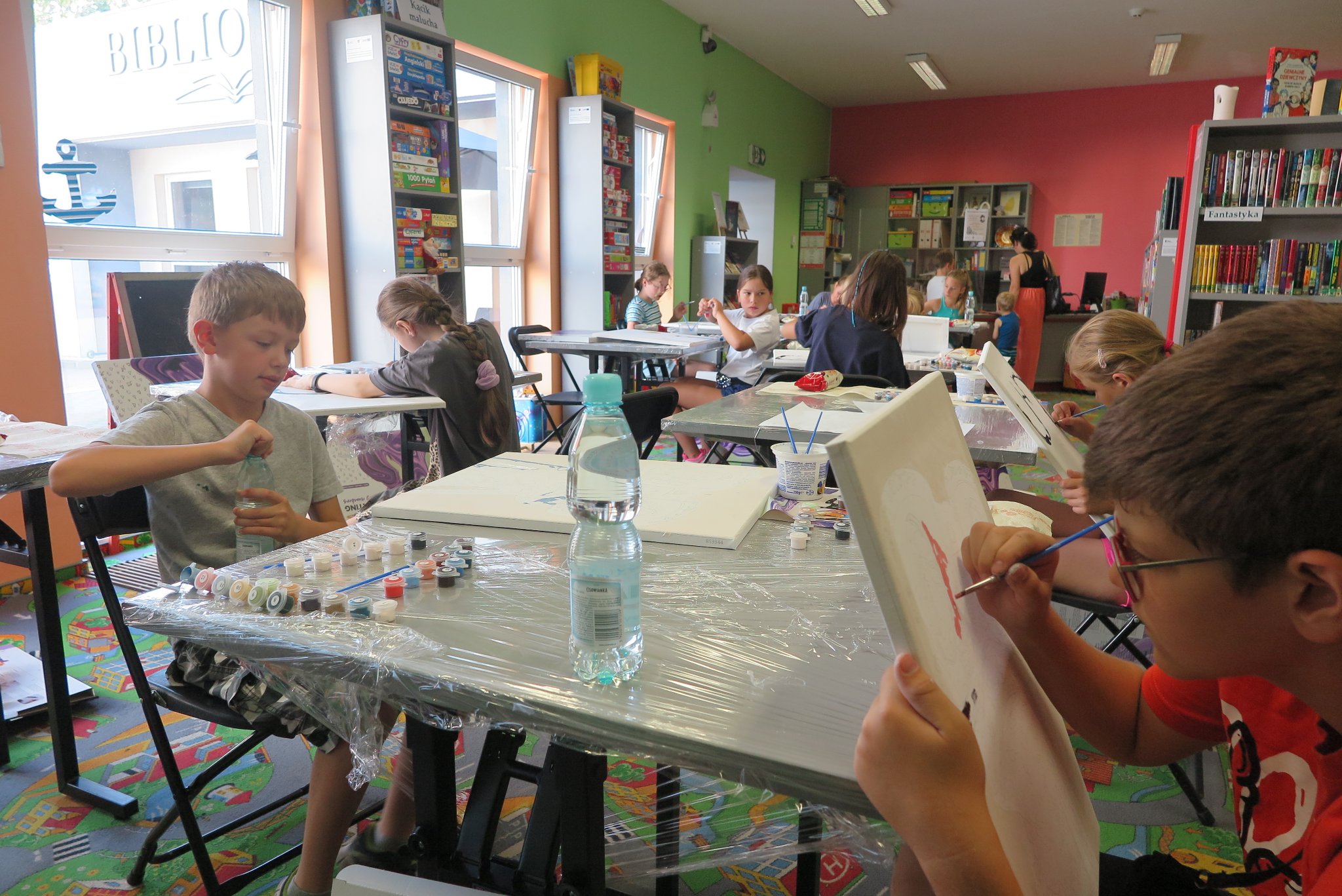 uczestnicy warsztatów plastycznych malowania na płótnie, siedzą przy stolikach na których leżą płótna i farby, malują obrazy