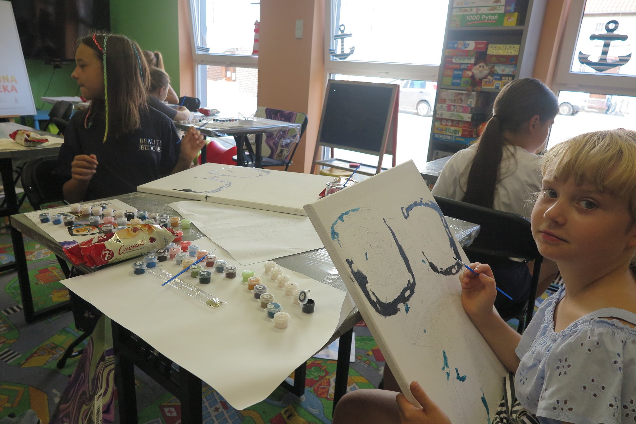 uczestnicy warsztatów plastycznych malowania na płótnie, siedzą przy stolikach na których leżą płótna i farby, malują obrazy