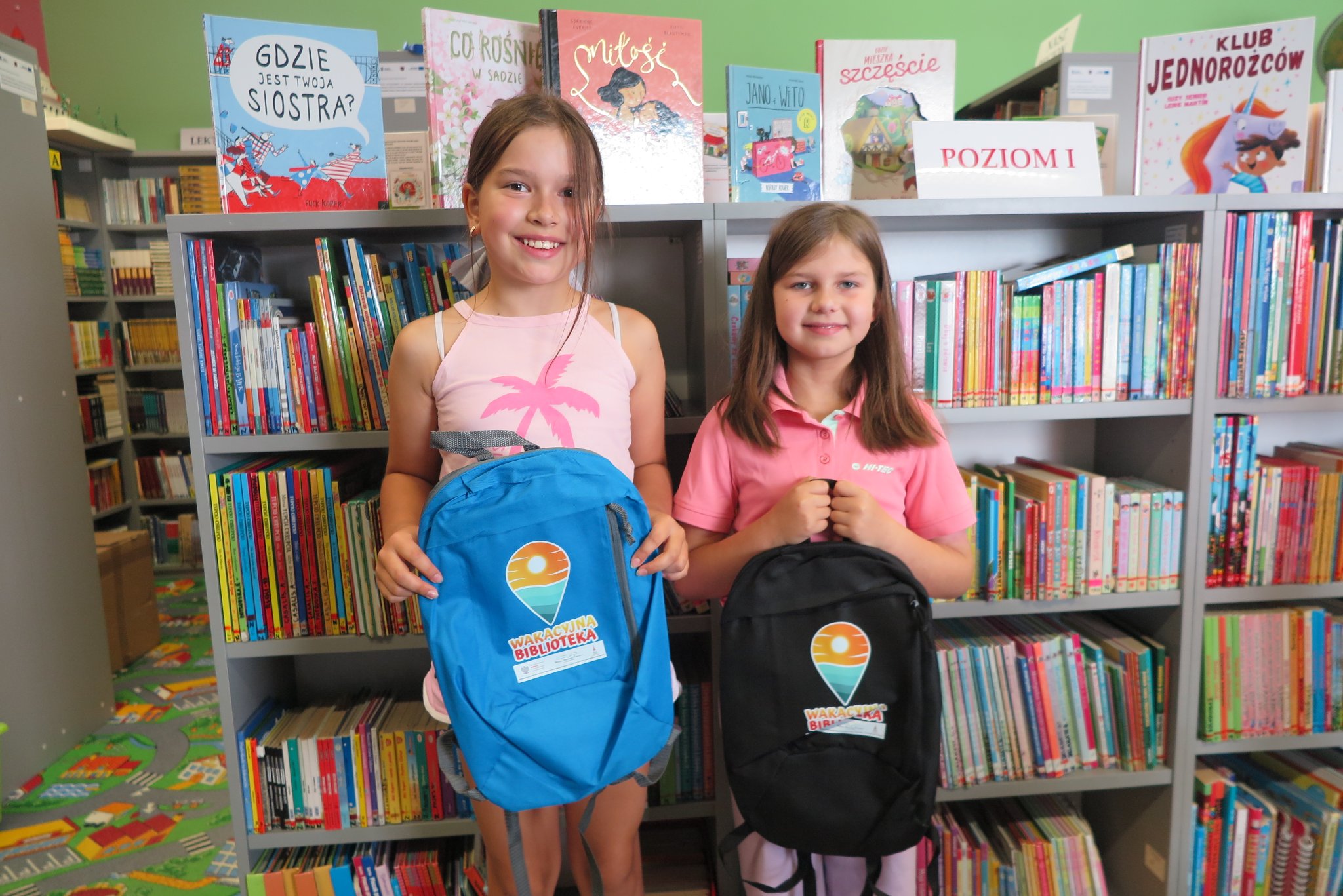 2 uczestniczki warsztatów z makramy stoją na tle regału z książkami, trzymają w rękach dwa plecaki z logo wakacyjnej biblioteki