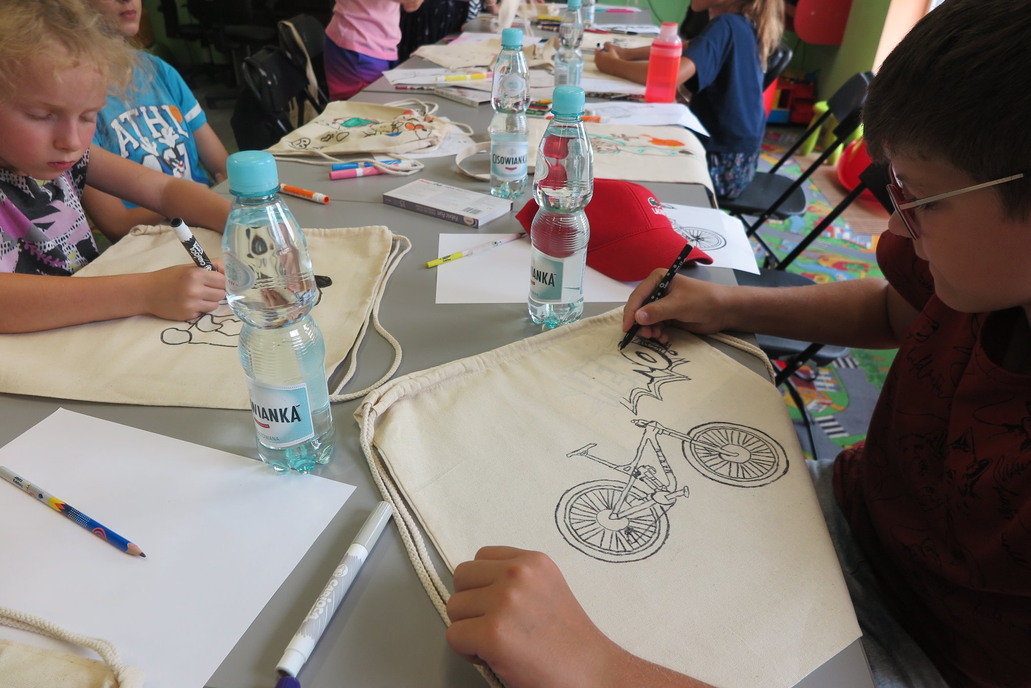 uczestnicy warsztatów z malowania bawełnianych toreb, siedzą przy długim stole i malują mazakami do tkanin białe torby