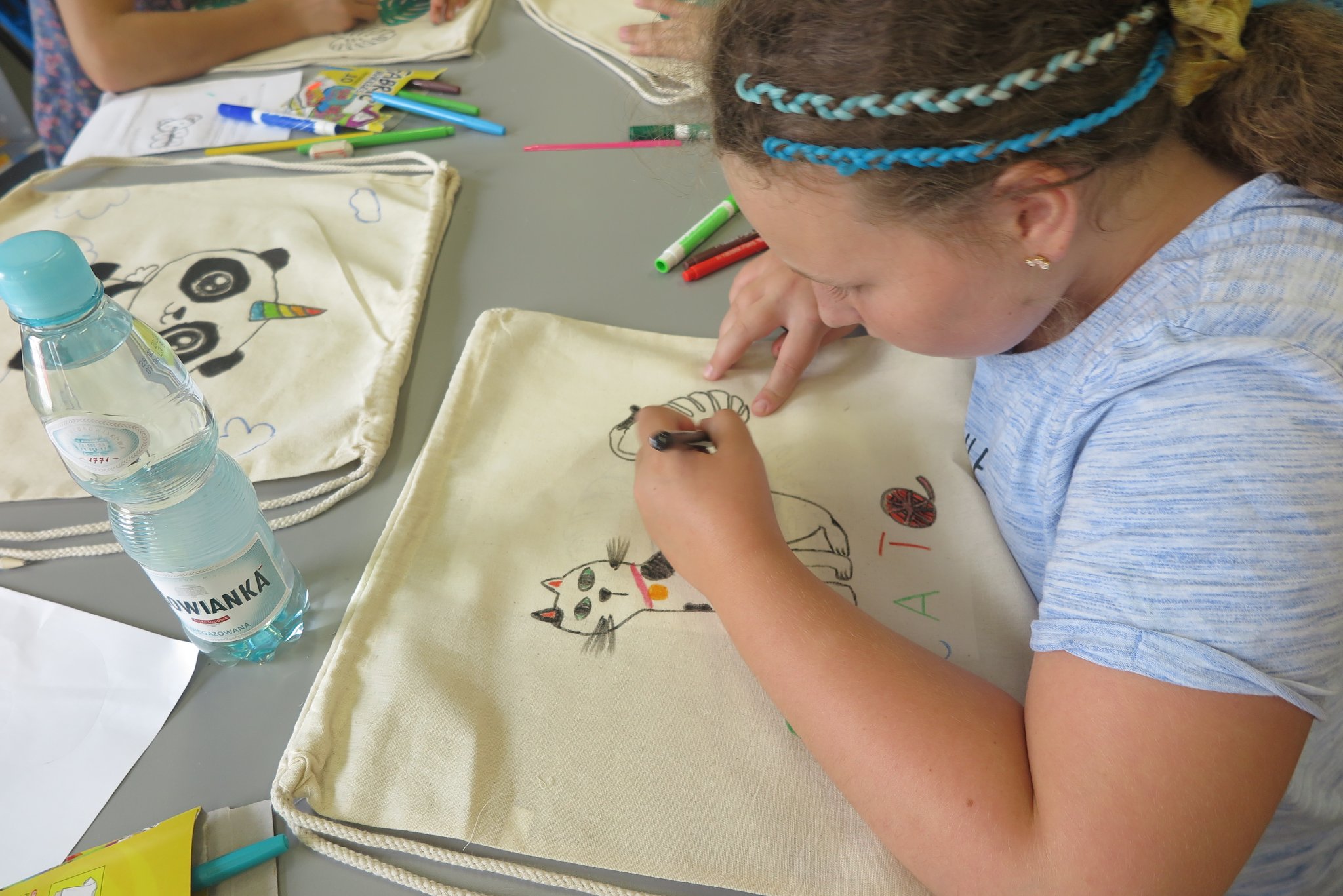 uczestnicy warsztatów z malowania bawełnianych toreb, siedzą przy długim stole i malują mazakami do tkanin białe torby
