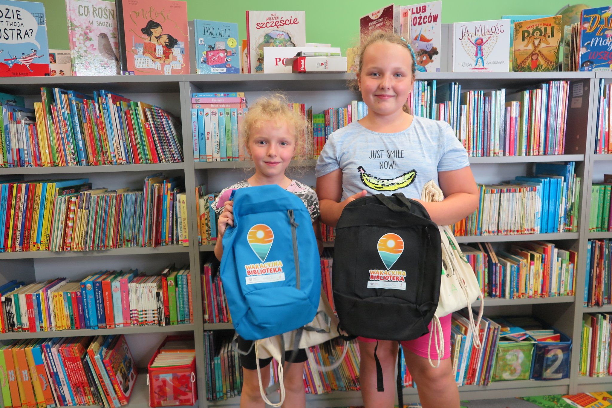 2 uczestniczki warsztatów z malowania toreb stoją na tle regału z książkami, trzymają w rękach dwa plecaki z logo wakacyjnej biblioteki