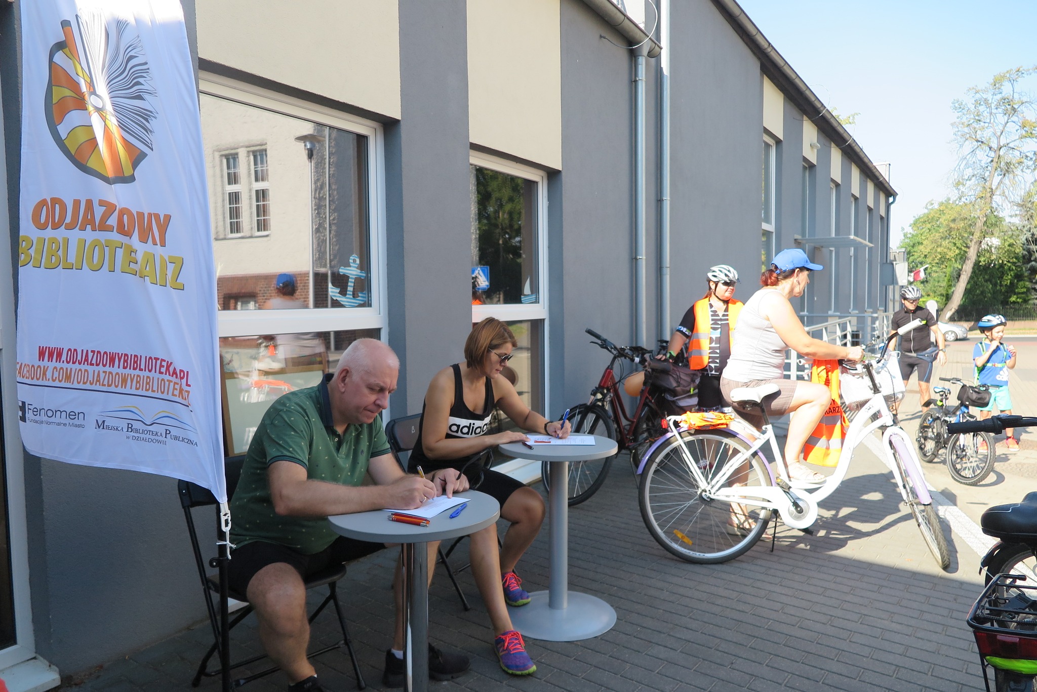 uczestnicy rajdu rowerowego zbierają się przed biblioteką, podpisują zgody na udział w rajdzie