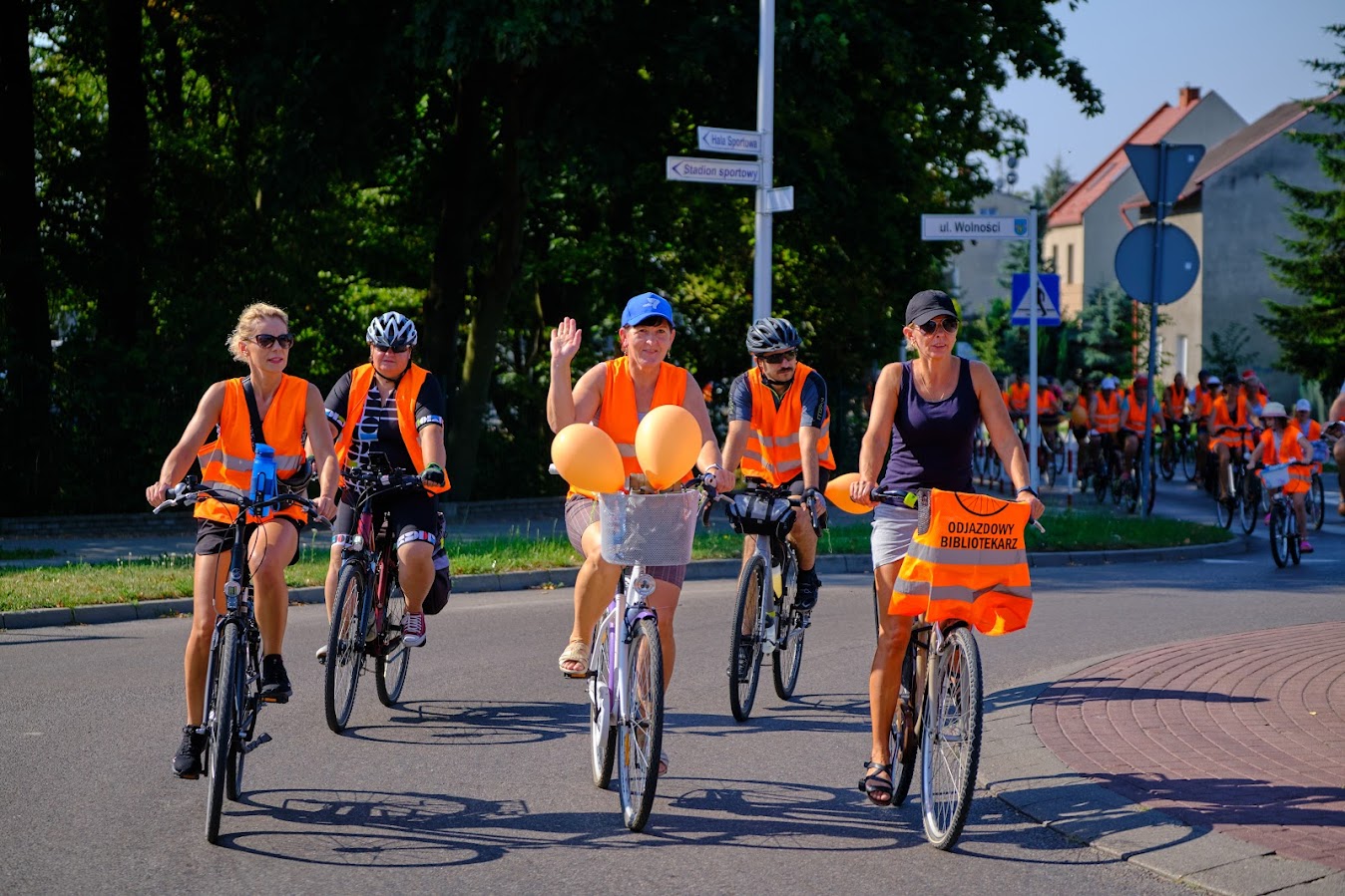 uczestnicy rajdu rowerowego jadą drogą publiczną (rondo), wszyscy mają założone pomarańczowe kamizelki odblaskowe z napisem Odjazdowy Bibliotekarz