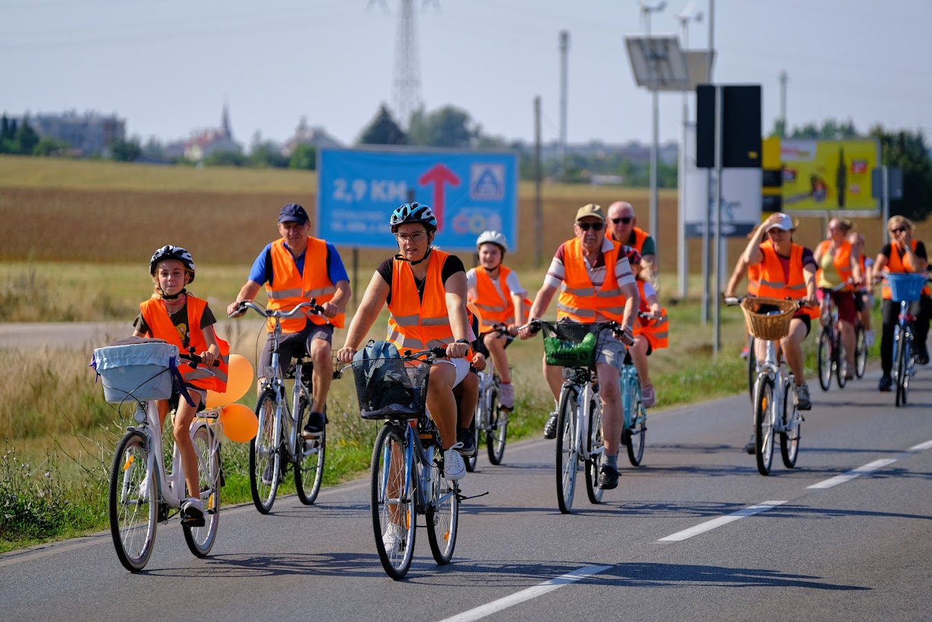 uczestnicy rajdu rowerowego jadą drogą publiczną, wszyscy mają założone pomarańczowe kamizelki odblaskowe z napisem Odjazdowy bibliotekarz