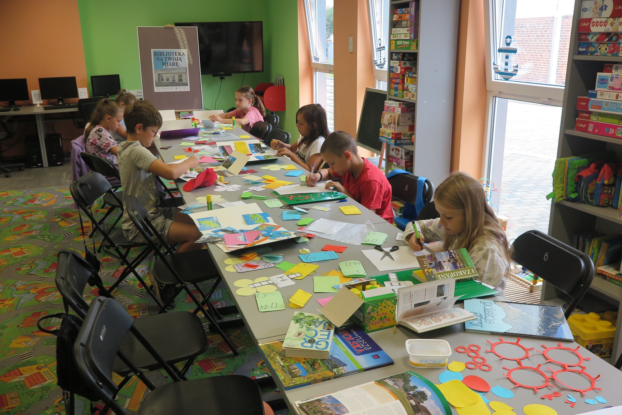 dzieci siedzą przy długim stole na którym leża materiały papiernicze, dzieci wykonują lapbooki 