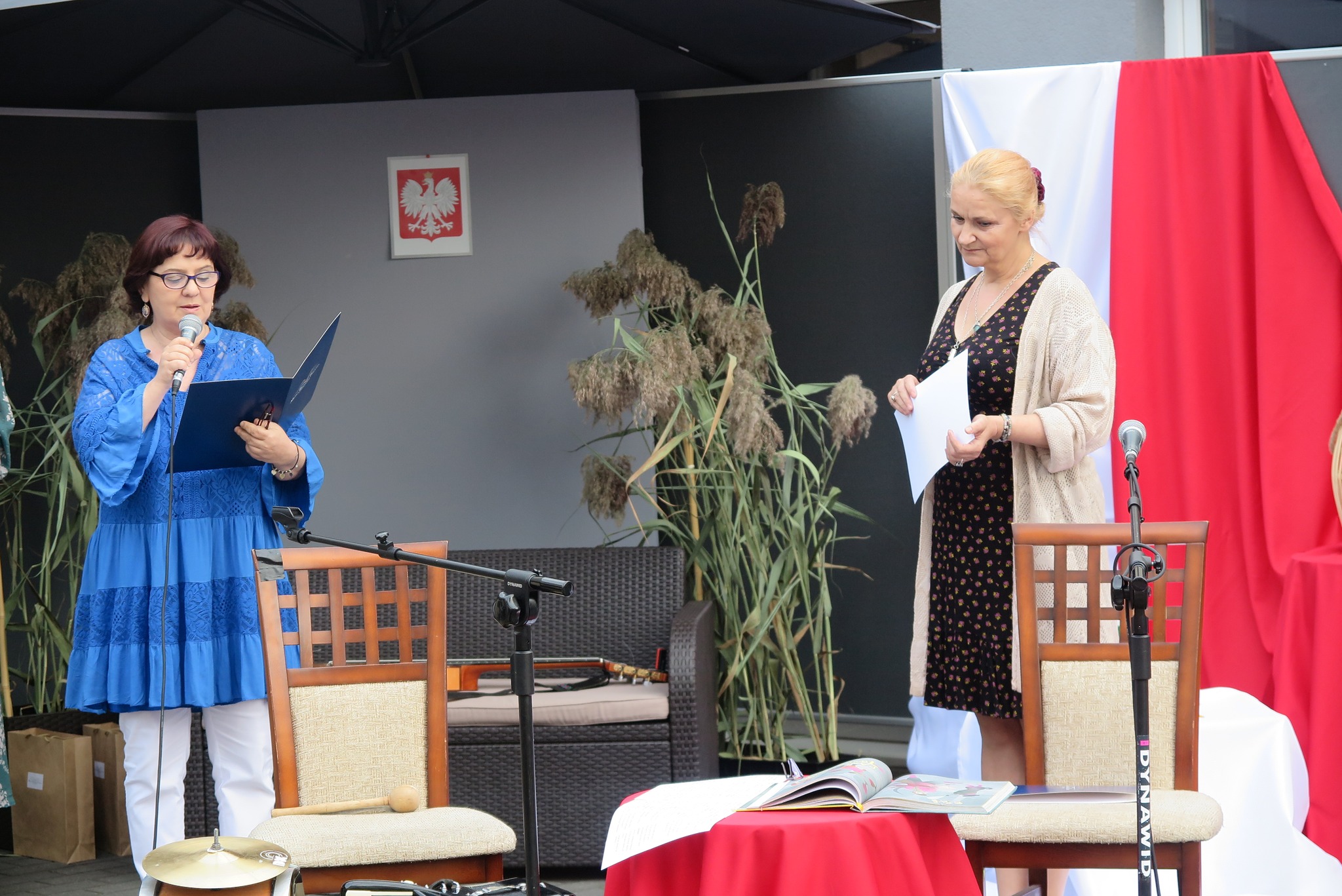 dwie kobiety stoją  przed publicznością, jedna z nich czyta do mikrofonu wiersz, druga trzyma kartkę