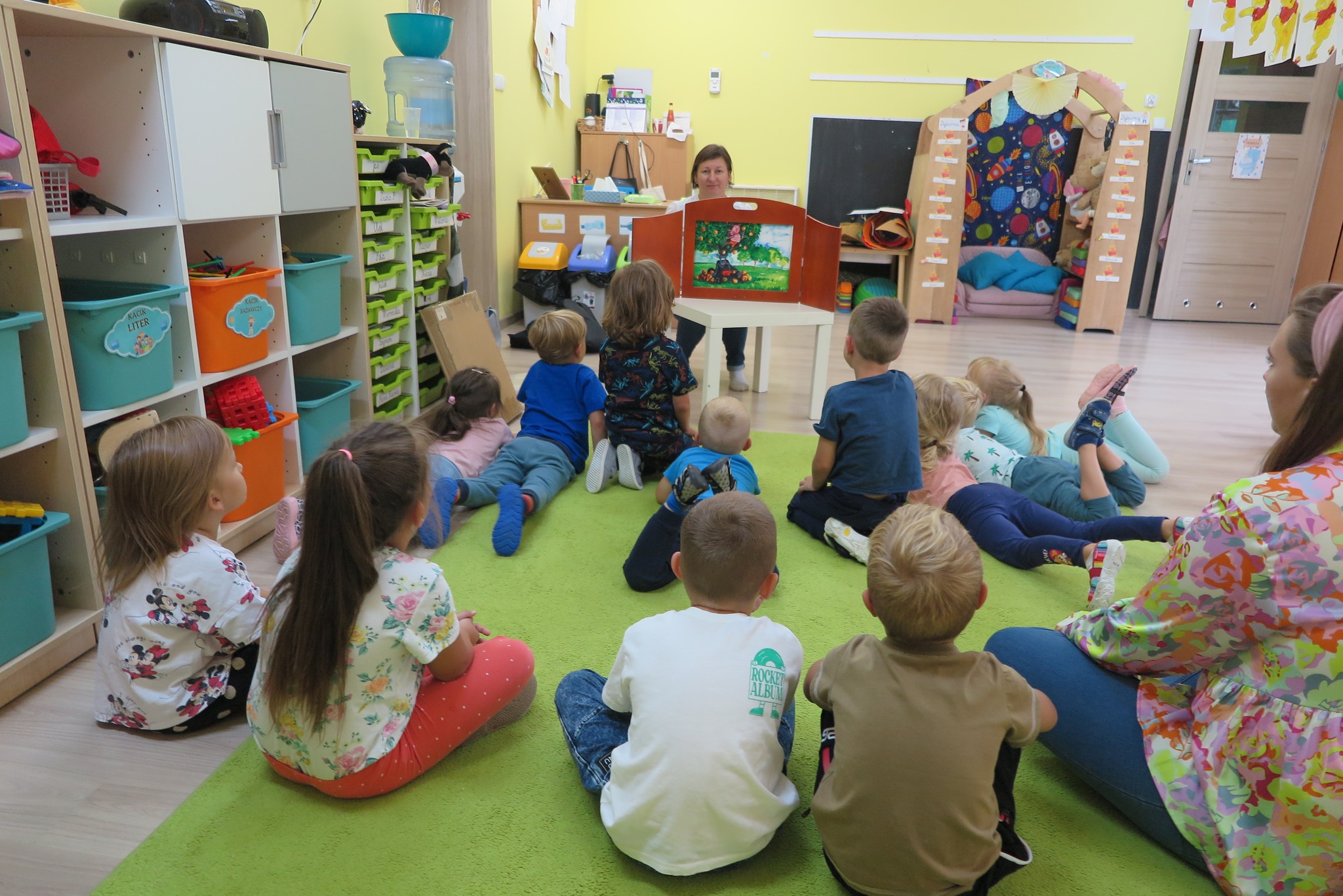 Na zdjęciu grupa dzieci siedząca na dywanie w przedszkolu. Razem z nimi na dywanie siedzi bibliotekarka. przed sobą ma rozłożony teatrzyk kamishibai