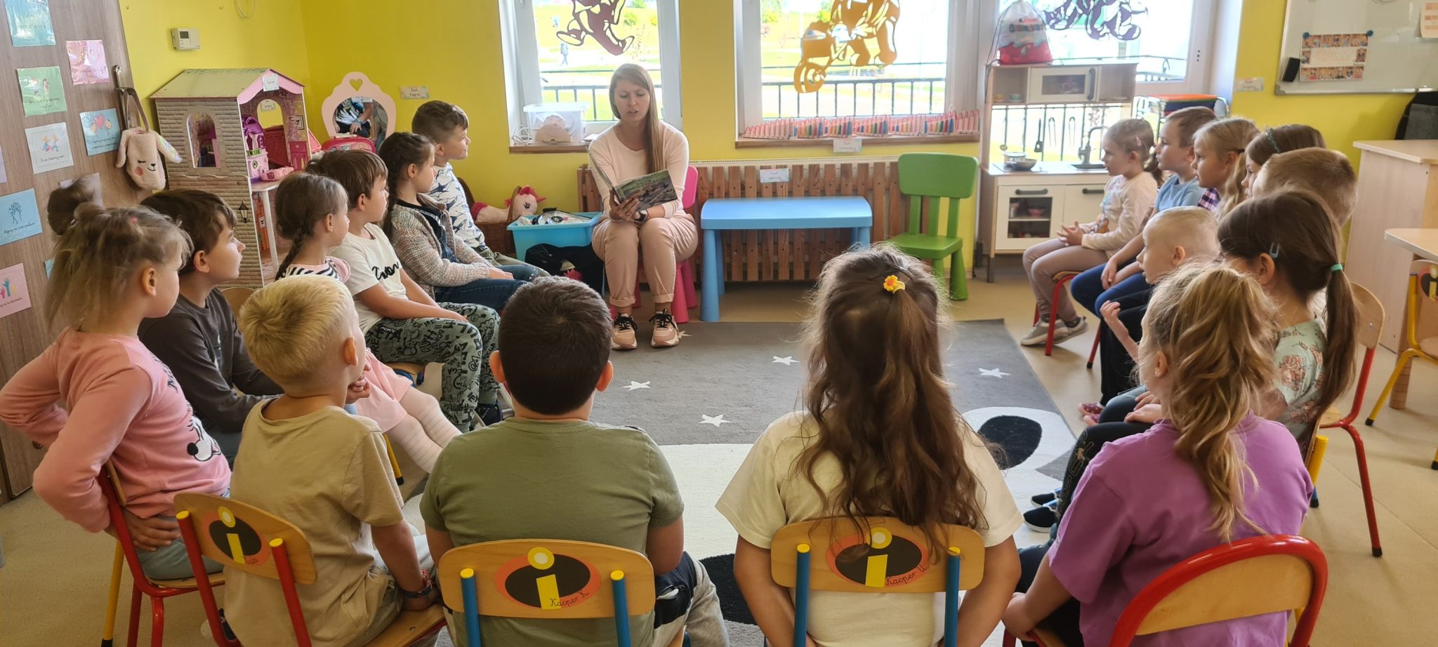 Na zdjęciu grupa dzieci siedząca krzesełkach w przedszkolu. Razem z nimi na krzesełku siedzi bibliotekarka. W dłoniach trzyma rozłożoną książkę, i czyta dzieciom bajkę
