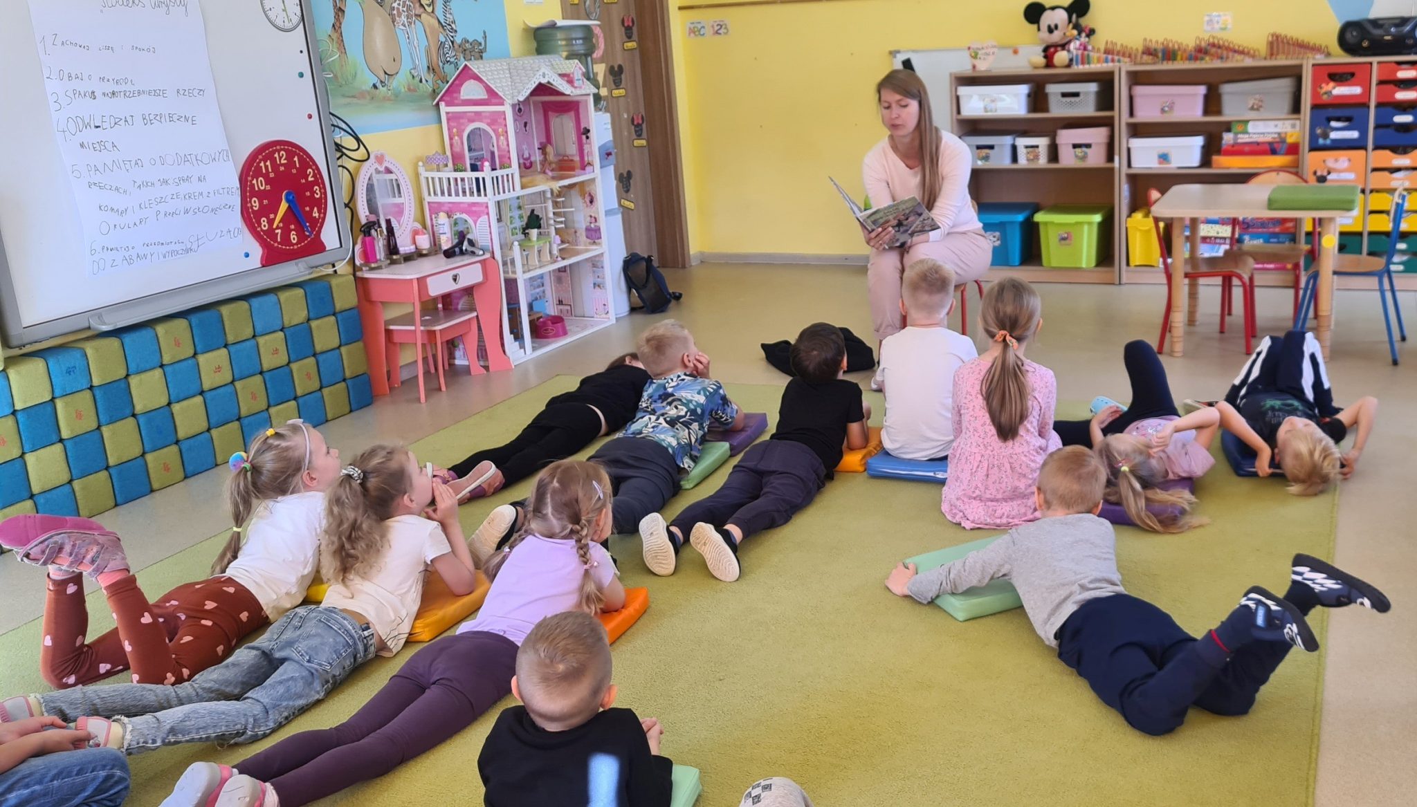 Na zdjęciu grupa dzieci leżąca na dywanie w przedszkolu. Razem z nimi na dywanie siedzi bibliotekarka. W dłoniach trzyma rozłożoną książkę, i czyta dzieciom bajkę