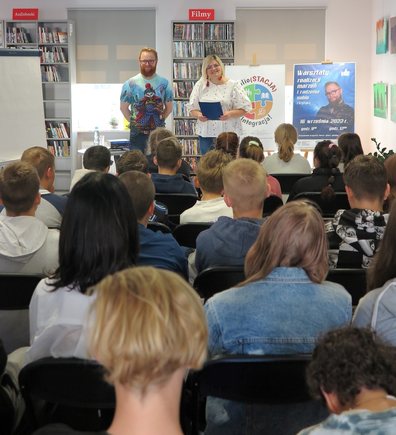 Krzysztof Piersa i bibliotekarka stoją przed publicznością, za nim po lewej stoi flipchart a po prawej na sztaludze dwa plakaty, logo Biblio(STACJA) - Integracja! i warsztaty realizacji marzeń i radzenia sobie z krytyką