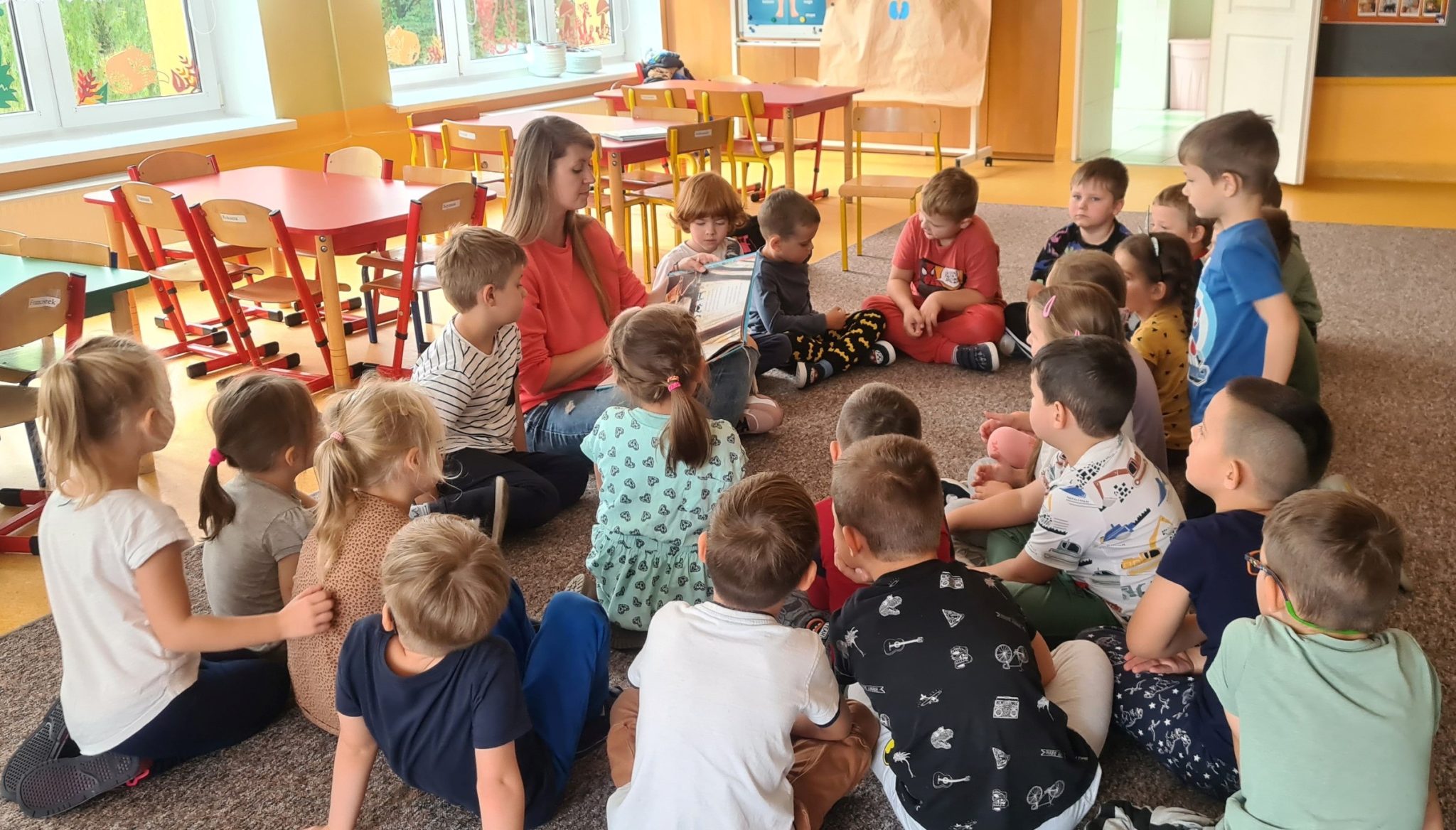 Na zdjęciu grupa dzieci siedząca na dywanie w przedszkolu. Razem z nimi na dywanie siedzi bibliotekarka. W dłoniach trzyma rozłożoną książkę, i czyta dzieciom bajkę