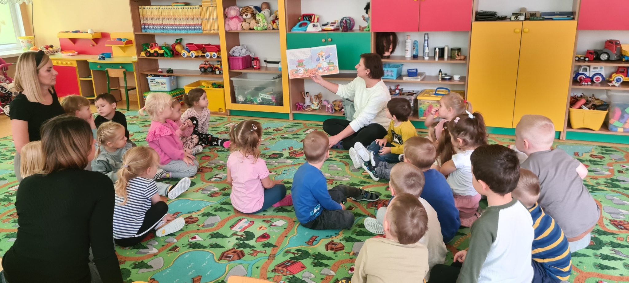 Na zdjęciu grupa dzieci siedząca na dywanie w przedszkolu. Razem z nimi na dywanie siedzi bibliotekarka. W dłoniach trzyma rozłożoną książkę, i czyta dzieciom bajkę