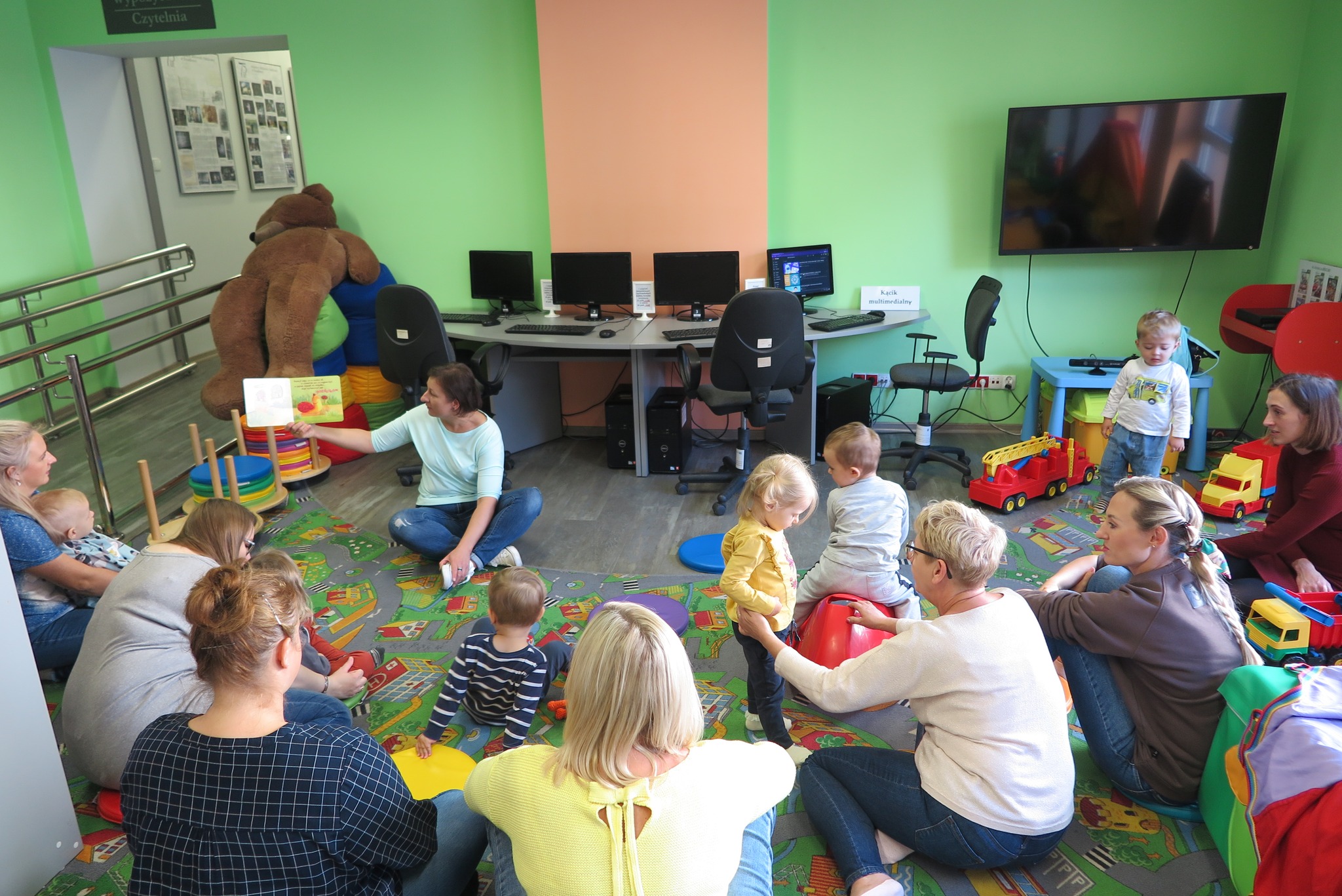 dzieci z rodziacami w oddziale dla dzieci siedzą na dywanie, bibliotekarka pokazuje książkę
