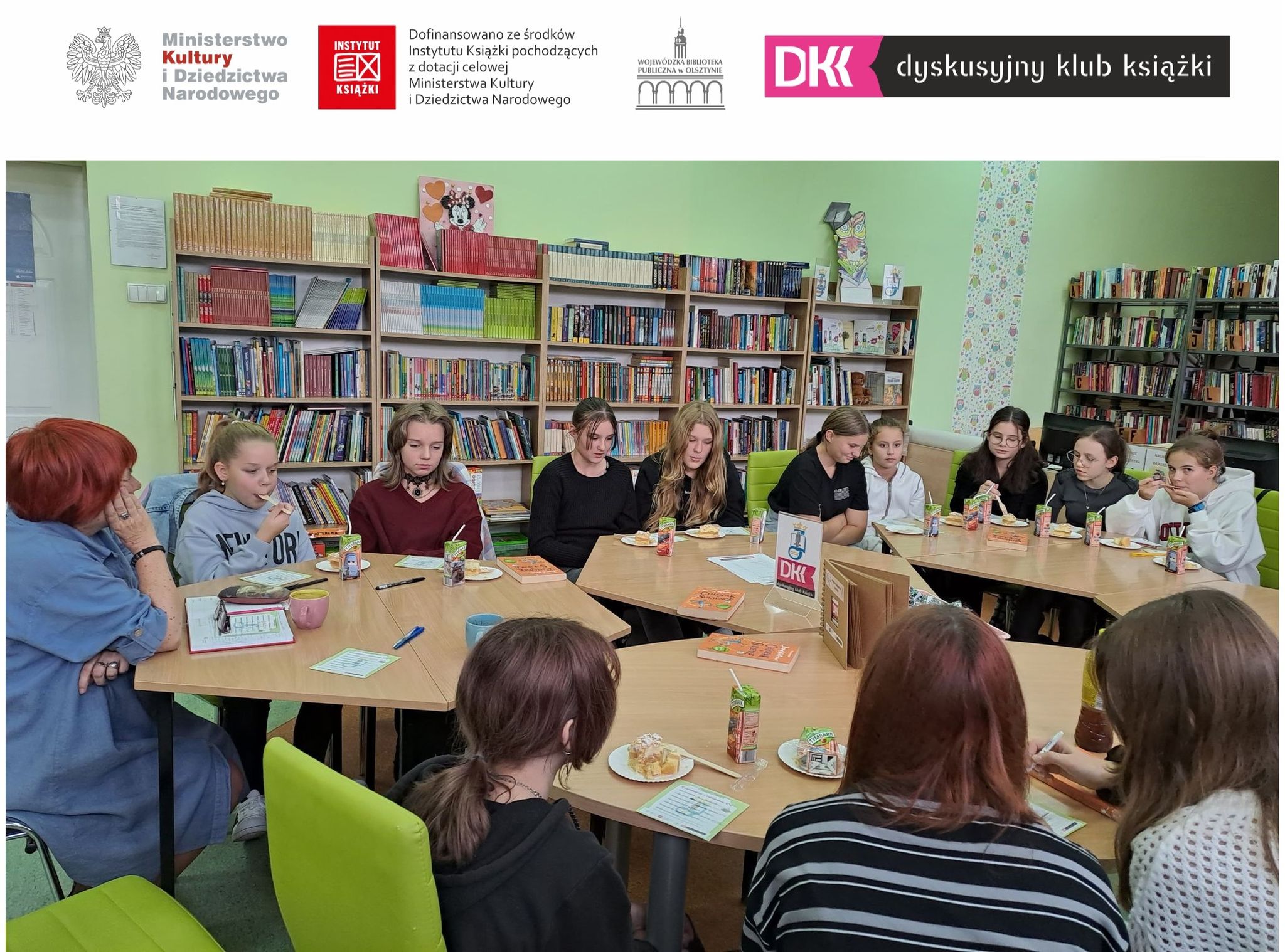 członkowie klubu dkk podczas spotkania w bibliotece szkolnej siedzą przy stolikach razem z osobą prowadzącą klub, na sole leżą książki i poczęstunek