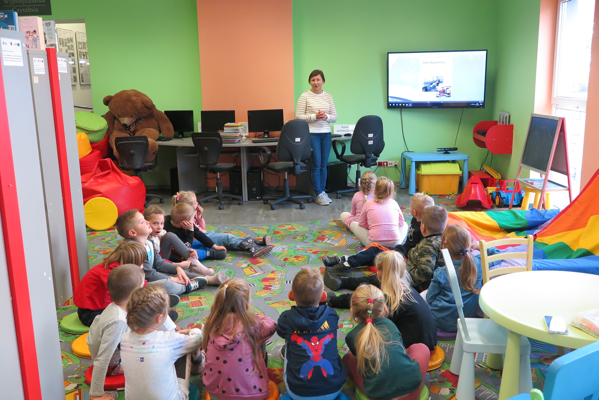grupa dzieci z przedszkola jest oprowadzana przez bibliotekarkę po oddziale dla dzieci, dzieci siedzą na dywanie w oddziale